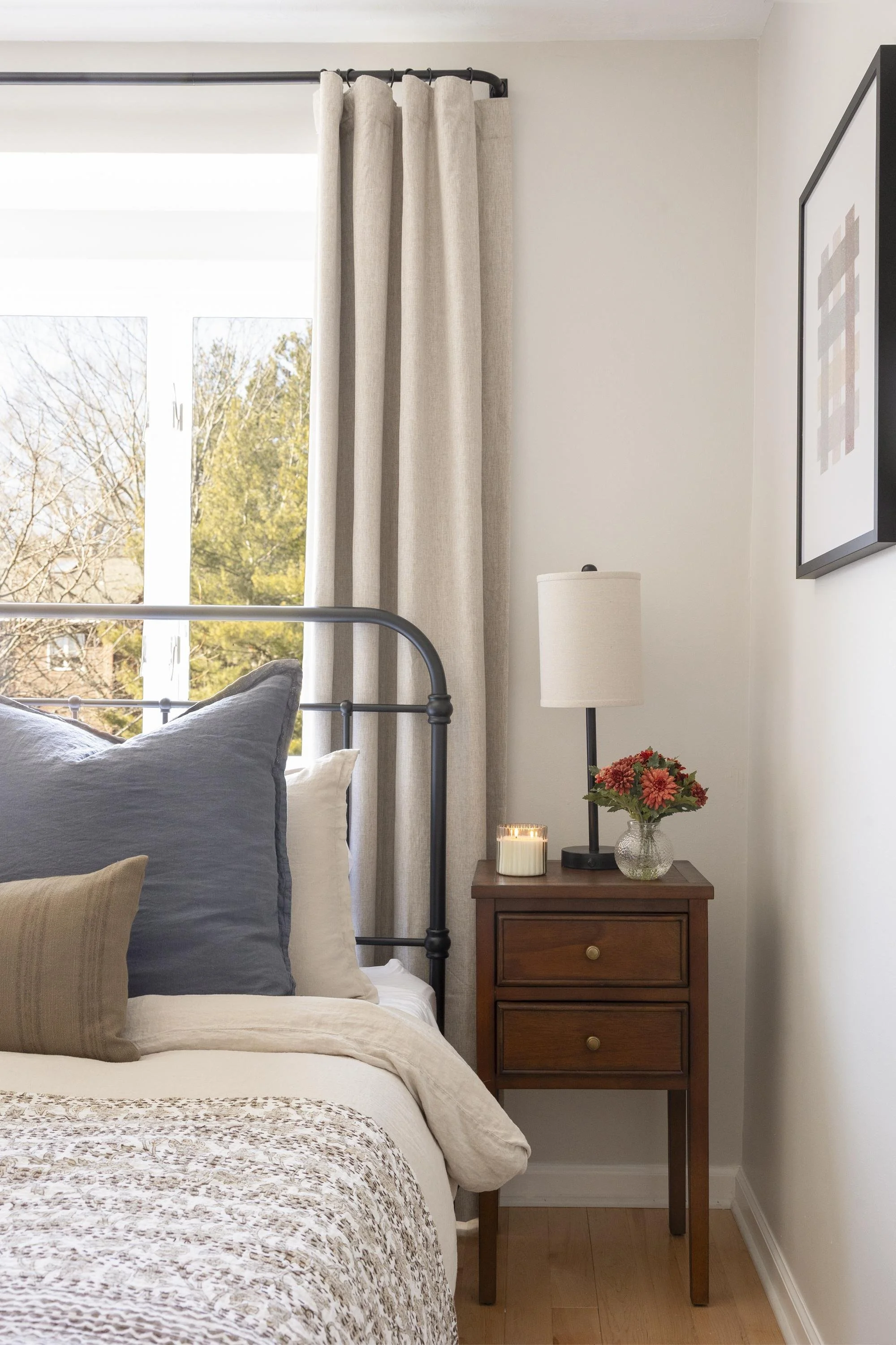 A cozy bedroom with a bed, gray and beige pillows, and a beige quilt. Next to the bed, a wooden nightstand holds a lit candle, a clear glass vase with pink and red flowers, and a black table lamp. Behind the bed, a window with beige curtains and sunl