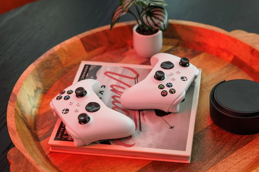 Two white Xbox controllers placed on top of a magazine on a wooden table. A potted plant is in the background and a black round object is on the right side of the table.