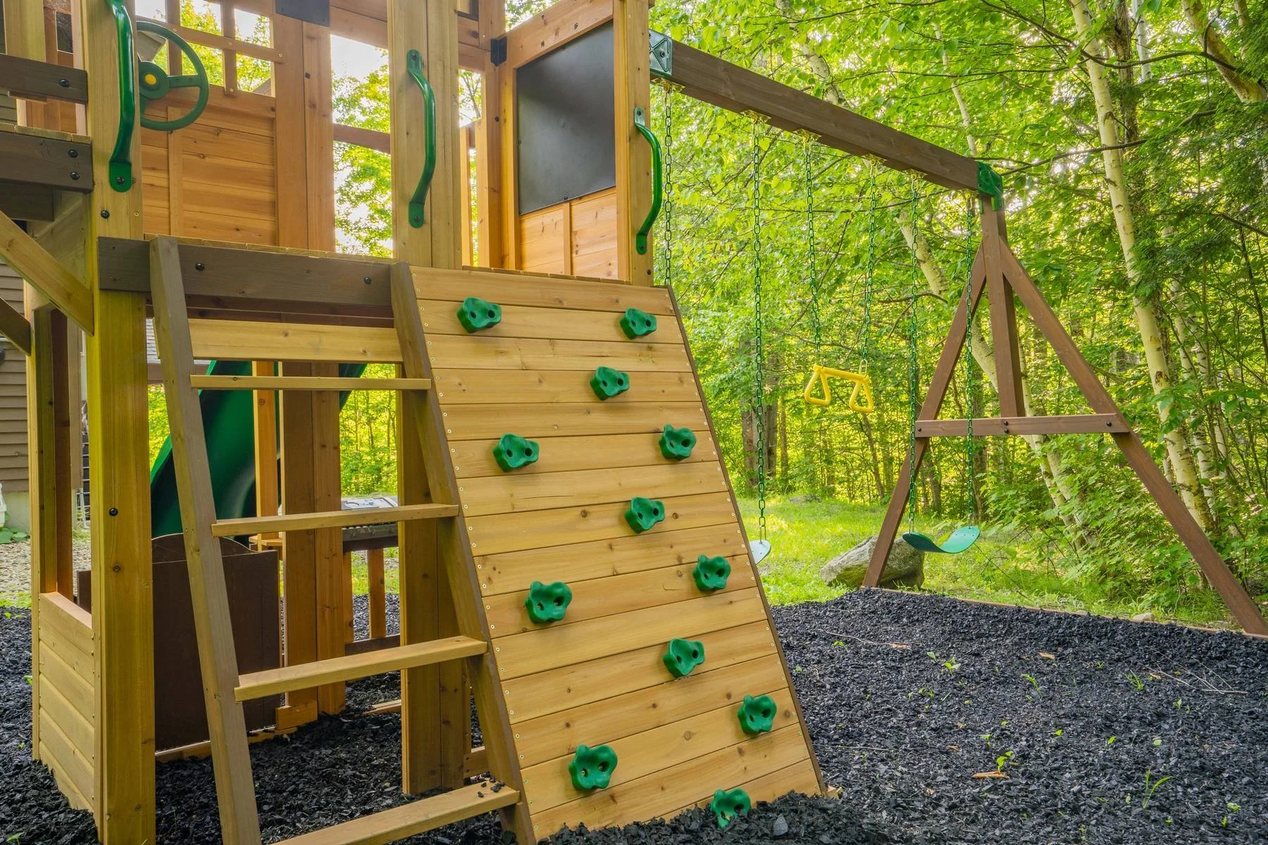 A wooden outdoor playground with a climbing wall, swing set, and slide, surrounded by trees and green foliage.