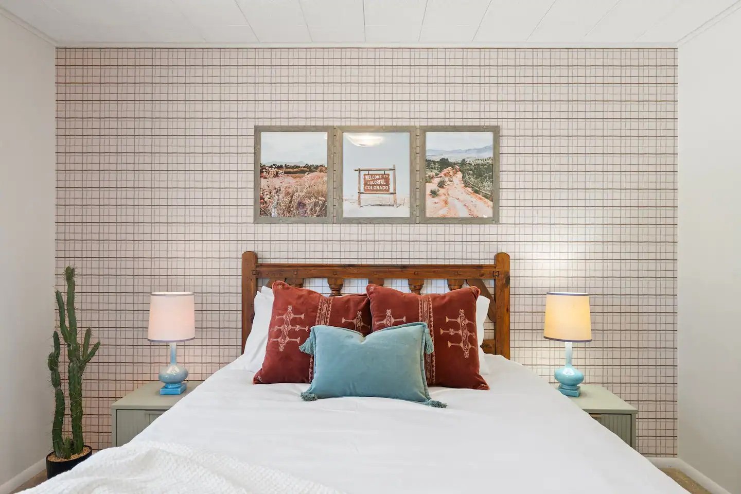 A cozy bedroom with a wooden bed frame, white bedding, and decorative pillows in red and teal. On each side of the bed are pale blue lamps, with a tall cactus in a black pot on the left. The back wall features a grid-patterned wallpaper, and above th