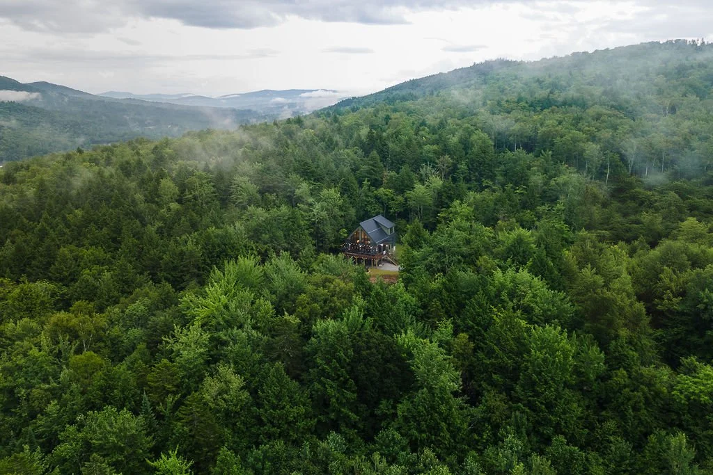 A house situated on a mountain slope surrounded by a dense forest of green trees, with mountains in the background and a cloudy sky.