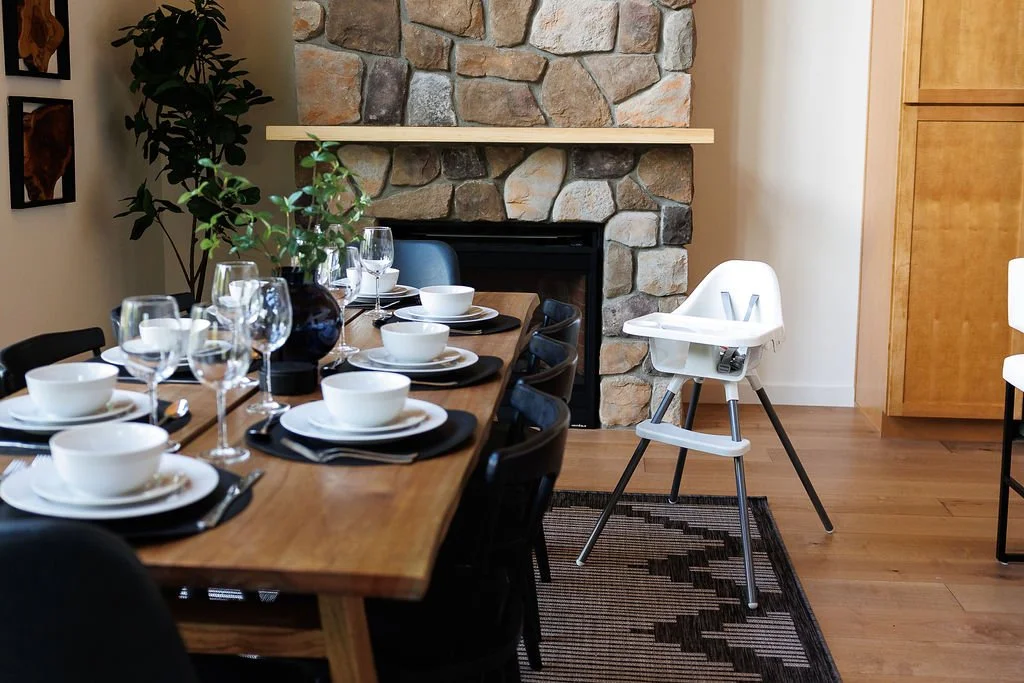 A dining room with a wooden table set with plates, bowls, utensils, and wine glasses. A high chair is positioned near the table. The room features a stone fireplace, wooden flooring, a plant, and some wall art.