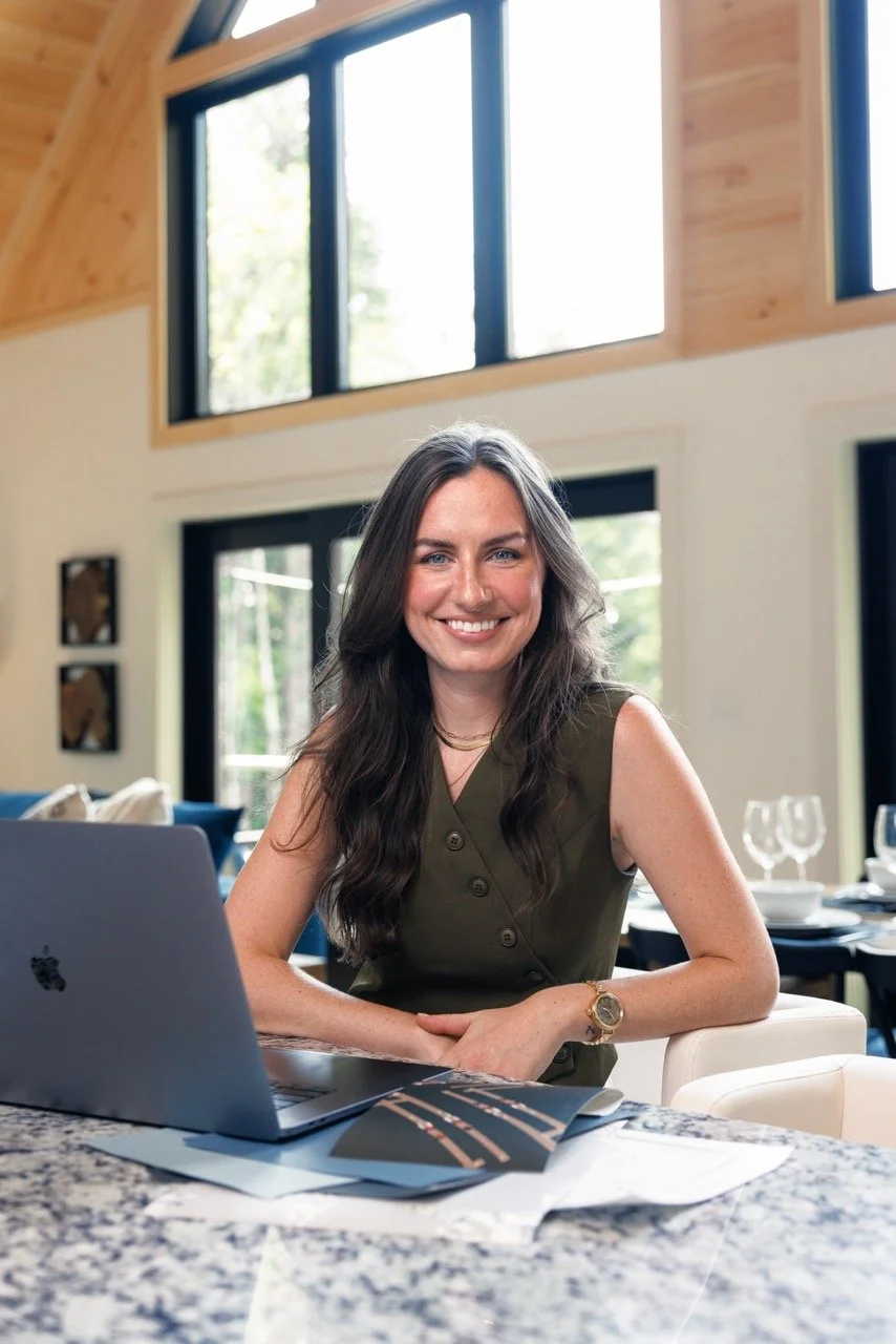 A smiling woman with long dark hair sitting at a granite countertop, working on a laptop in a modern, well-lit home or office space with large windows and wooden accents.