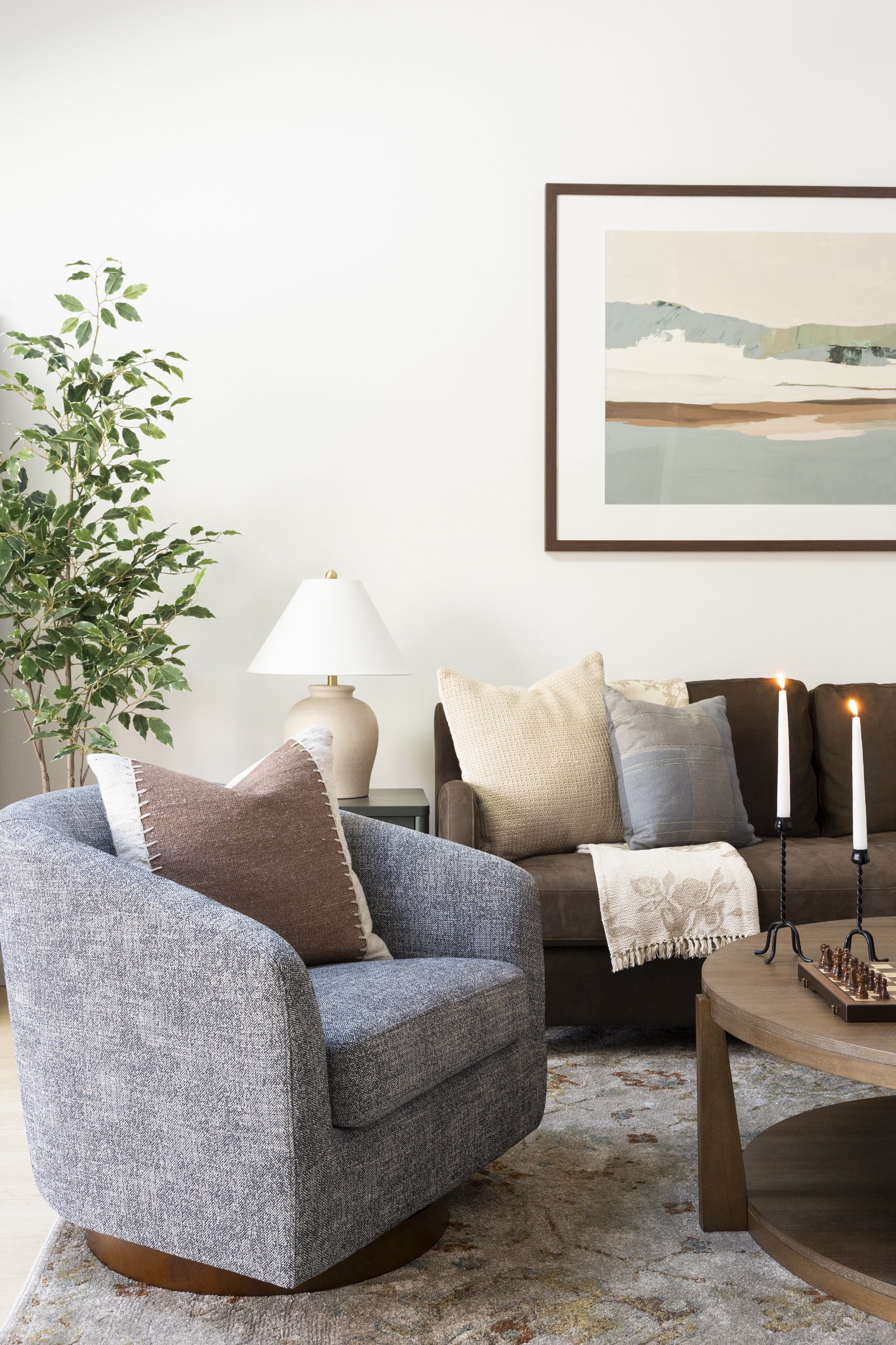 Living room with gray armchair in the foreground, brown sofa with beige and gray cushions, a side table with a white lamp, a framed landscape painting on the wall, a green plant on the left, a coffee table with candles and a chessboard, and a rug wit