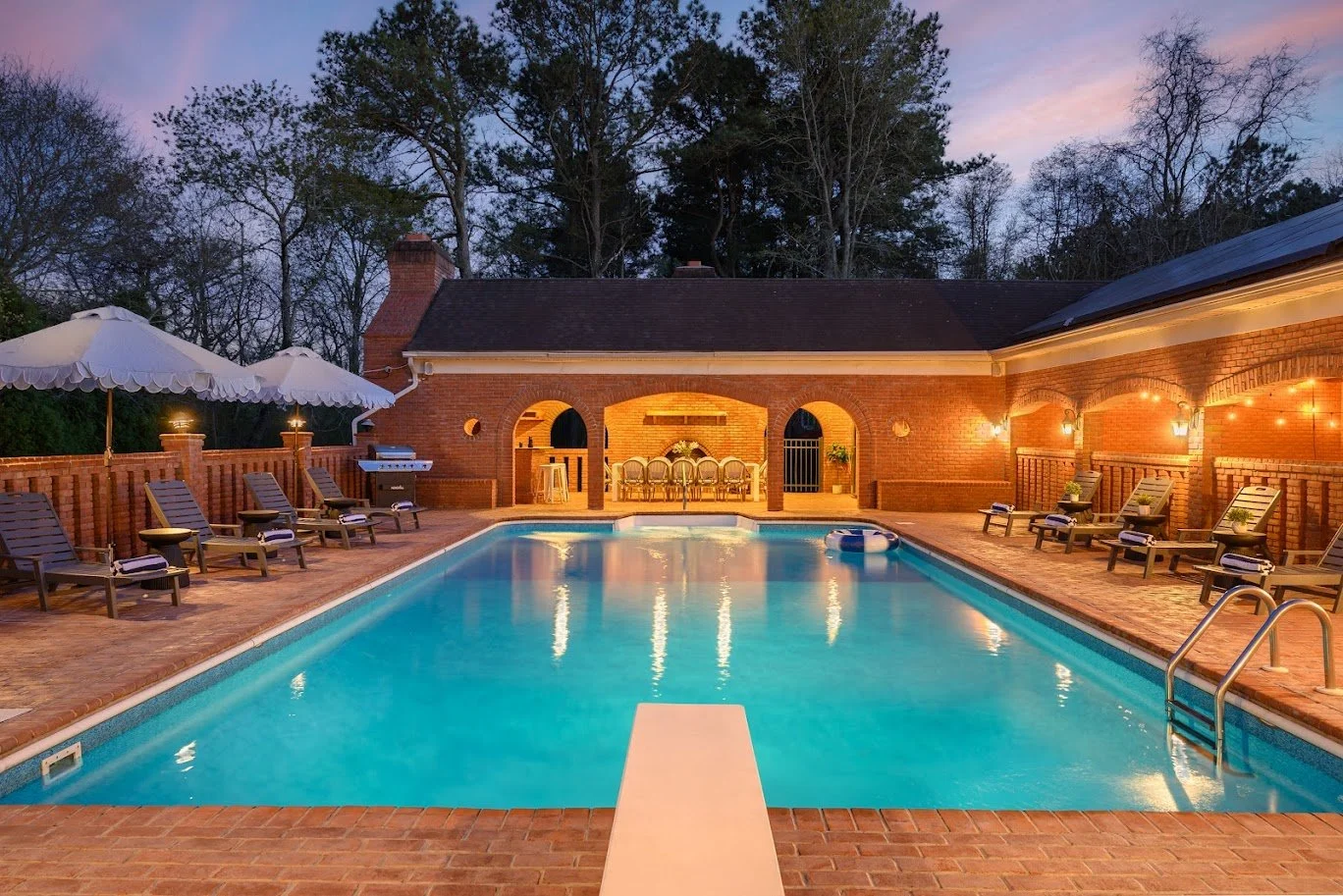 Residential backyard swimming pool with lounge chairs, umbrellas, brick patio, and a covered area with dining tables, illuminated by outdoor lights at dusk.