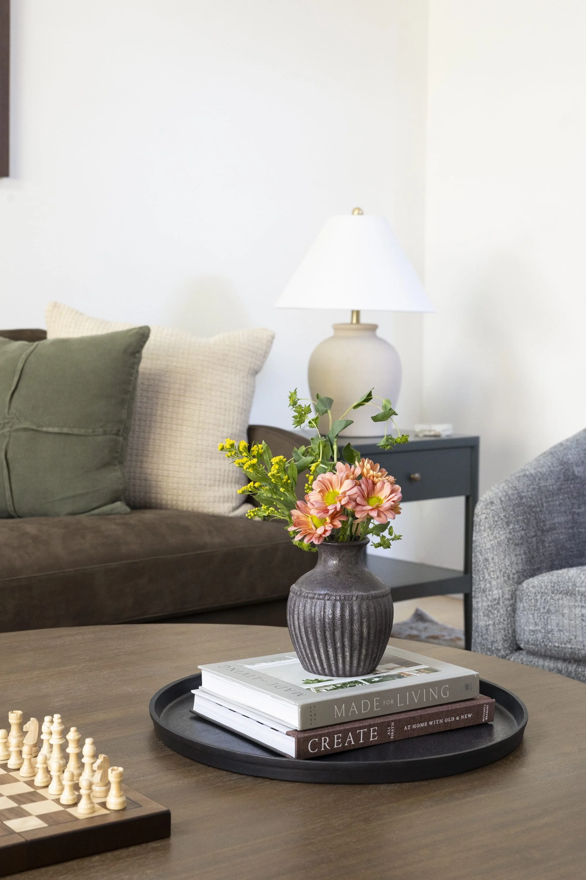 A living room with a wooden coffee table holding a black tray with stacked books labeled 'Made for Living,' 'Create,' and others, topped with a dark gray vase with pink and yellow flowers; a chessboard with pieces near the front left; a gray upholste