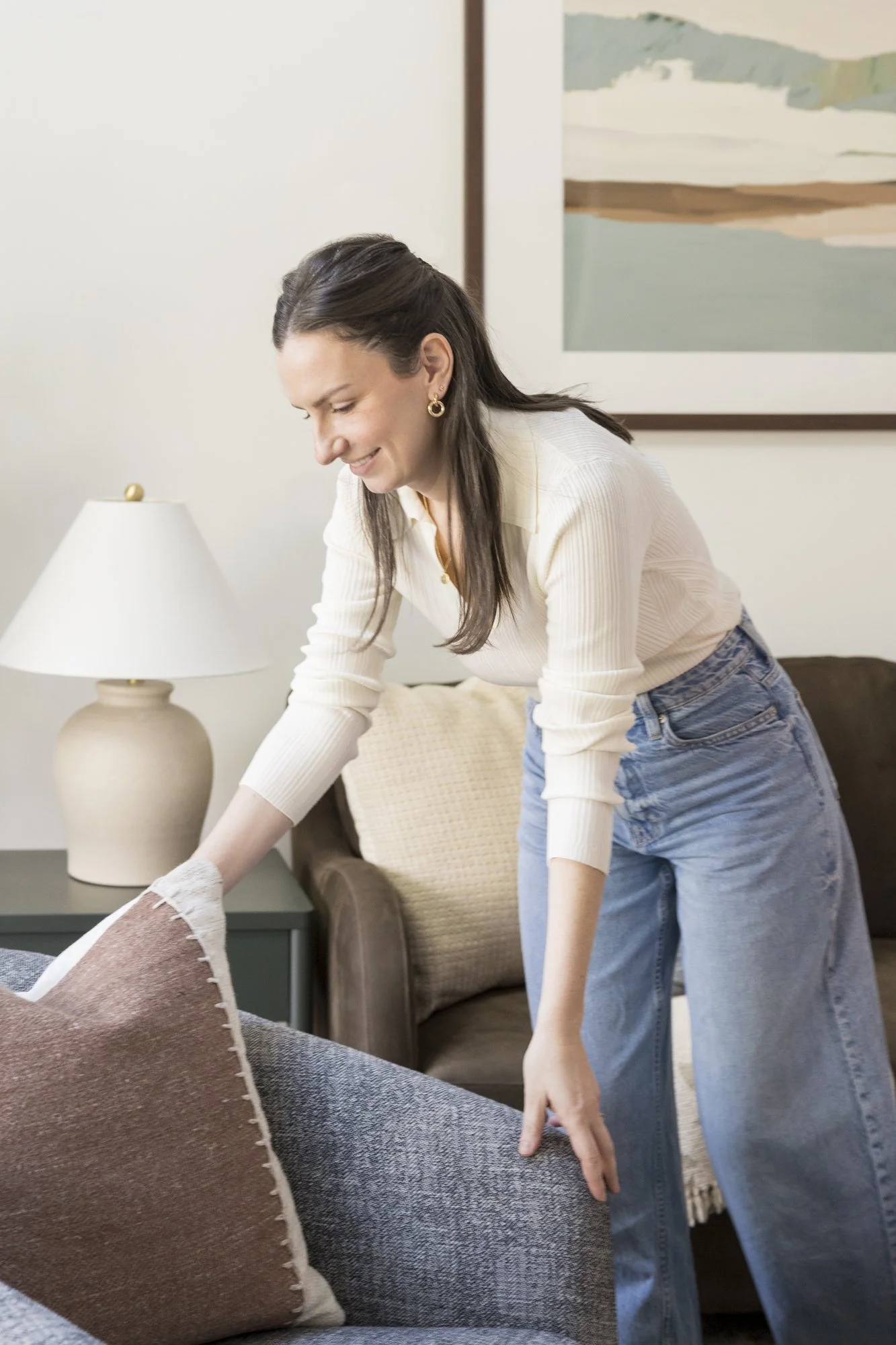 Woman cleaning a brown and gray throw pillow on a dark blue couch in a living room with a beige table lamp, a beige armchair with a pillow, and a large framed abstract landscape painting on the wall.