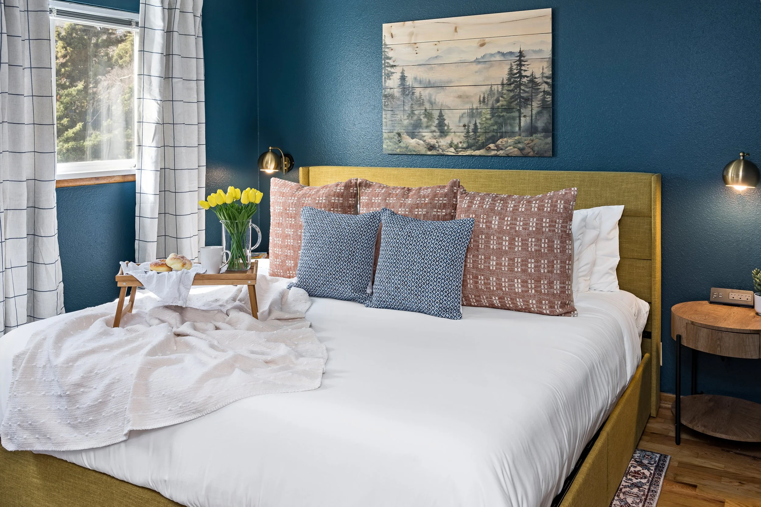 A cozy bedroom decorated with a yellow upholstered headboard and blue walls. There are multiple pillows on the bed, a tray with breakfast items including a vase of yellow tulips, and a window with white curtains. A landscape painting of pine trees an