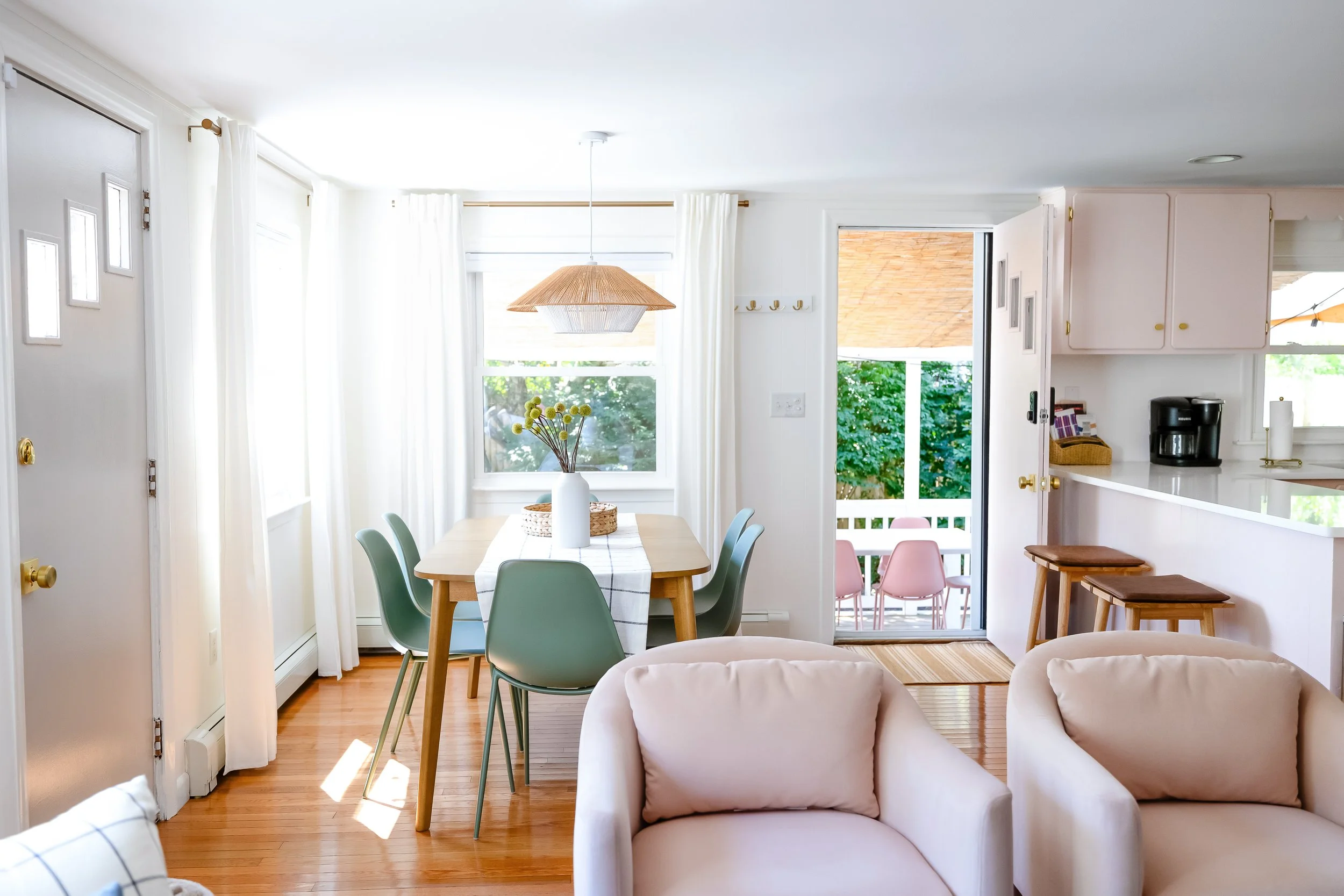 Bright dining area with a light wood table, pastel green chairs, and a white vase with yellow flowers, near large windows with white curtains, and a door leading to a balcony with trees outside.