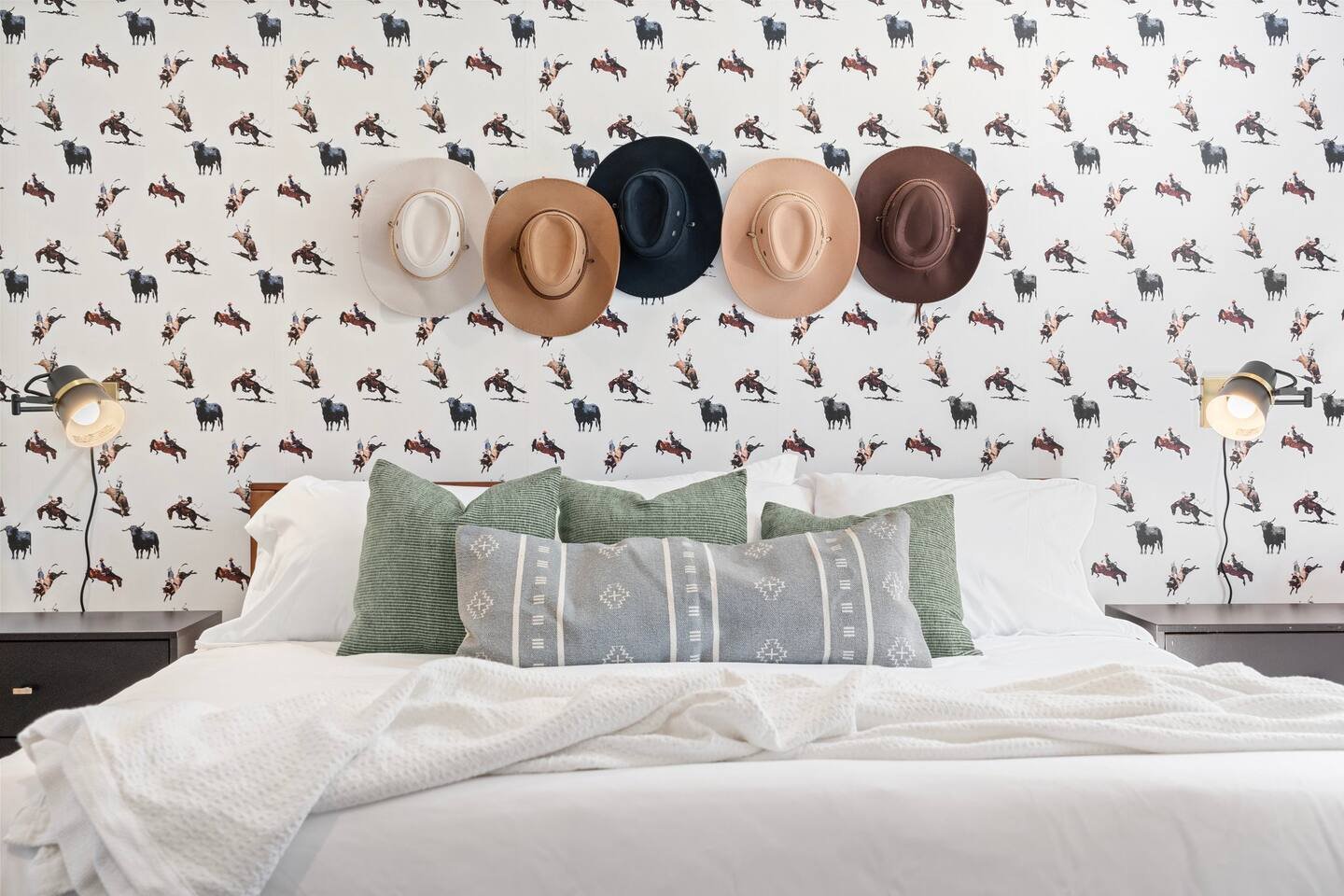 A bedroom with a bed, decorative pillows, wall-mounted lamps, and a patterned wallpaper featuring cowboys on horseback. Three hats hang on the wall above the bed.