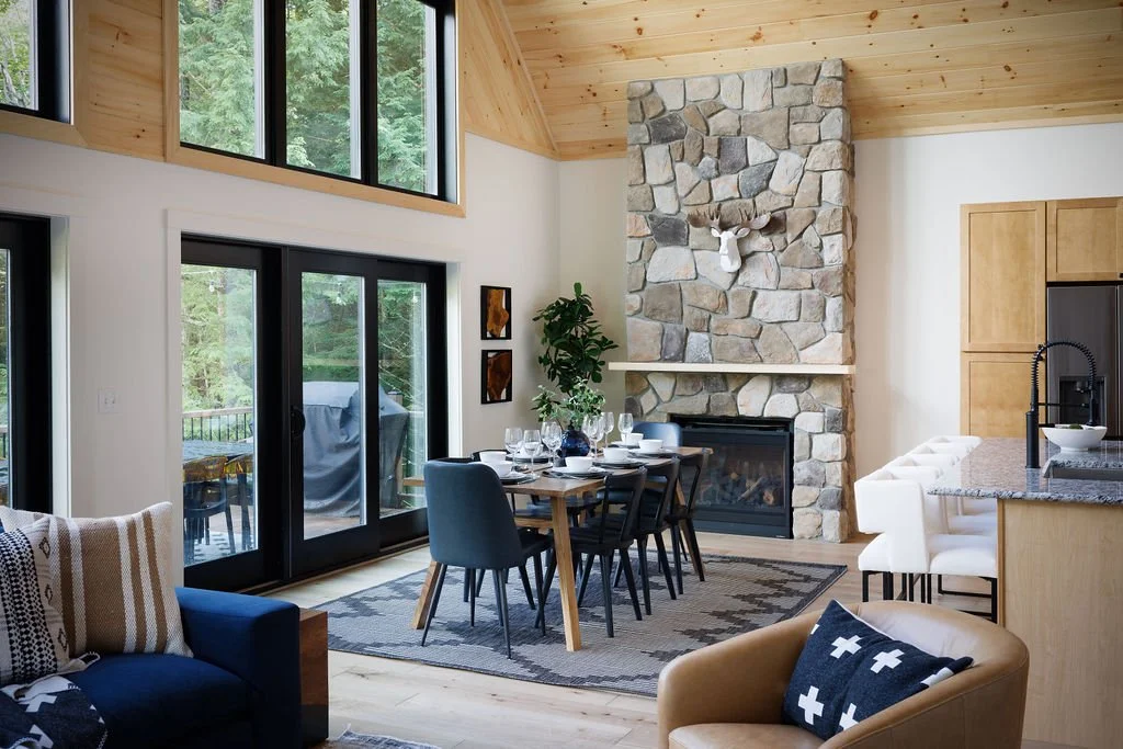 A living room with a stone fireplace, deer head mounted on it, a dining table set with glasses and plates, and large windows showing trees outside.