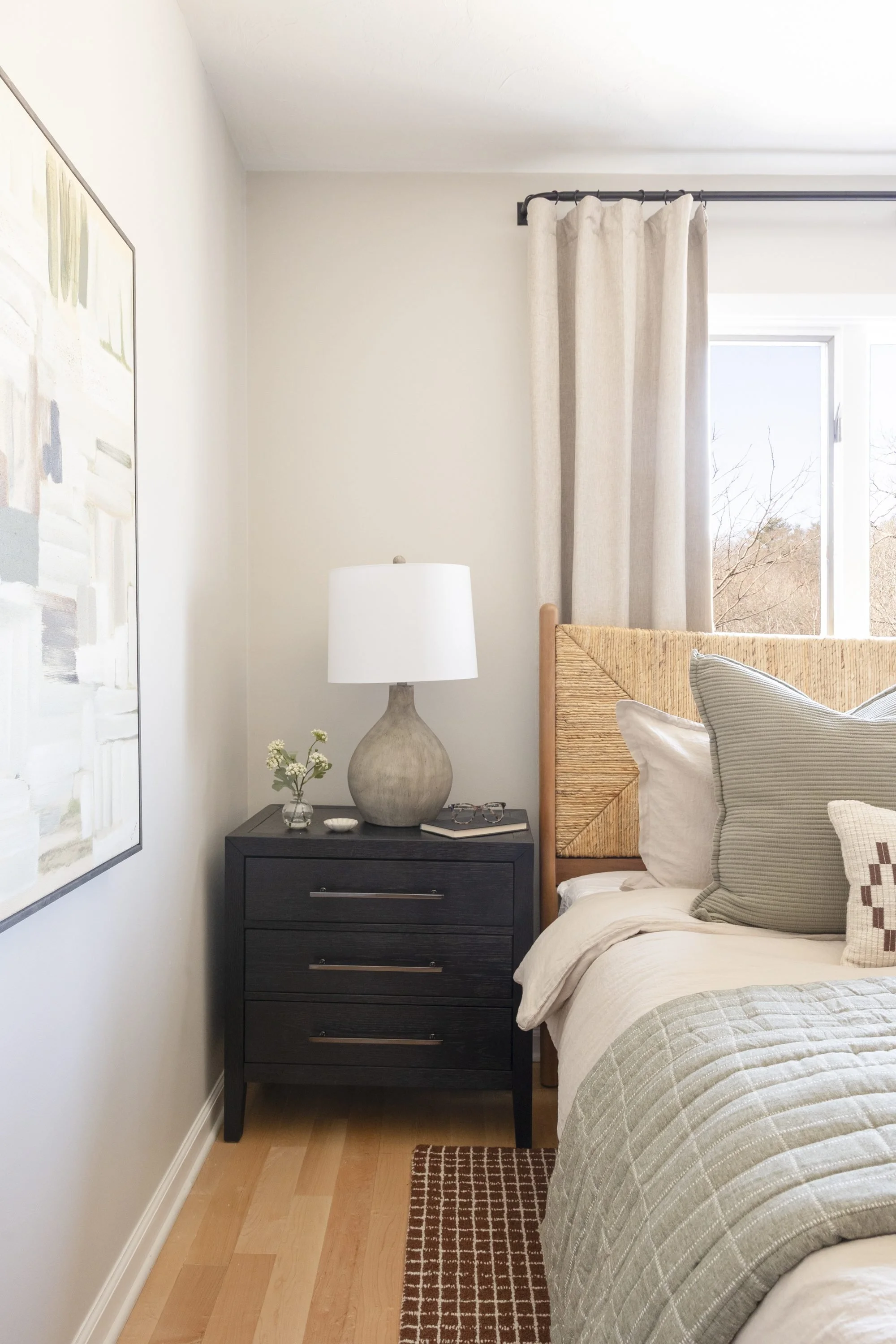 A bedroom corner with a bed, pillows, a black nightstand, a lamp, a small vase with flowers, a framed painting, curtains, and window with a view of trees.