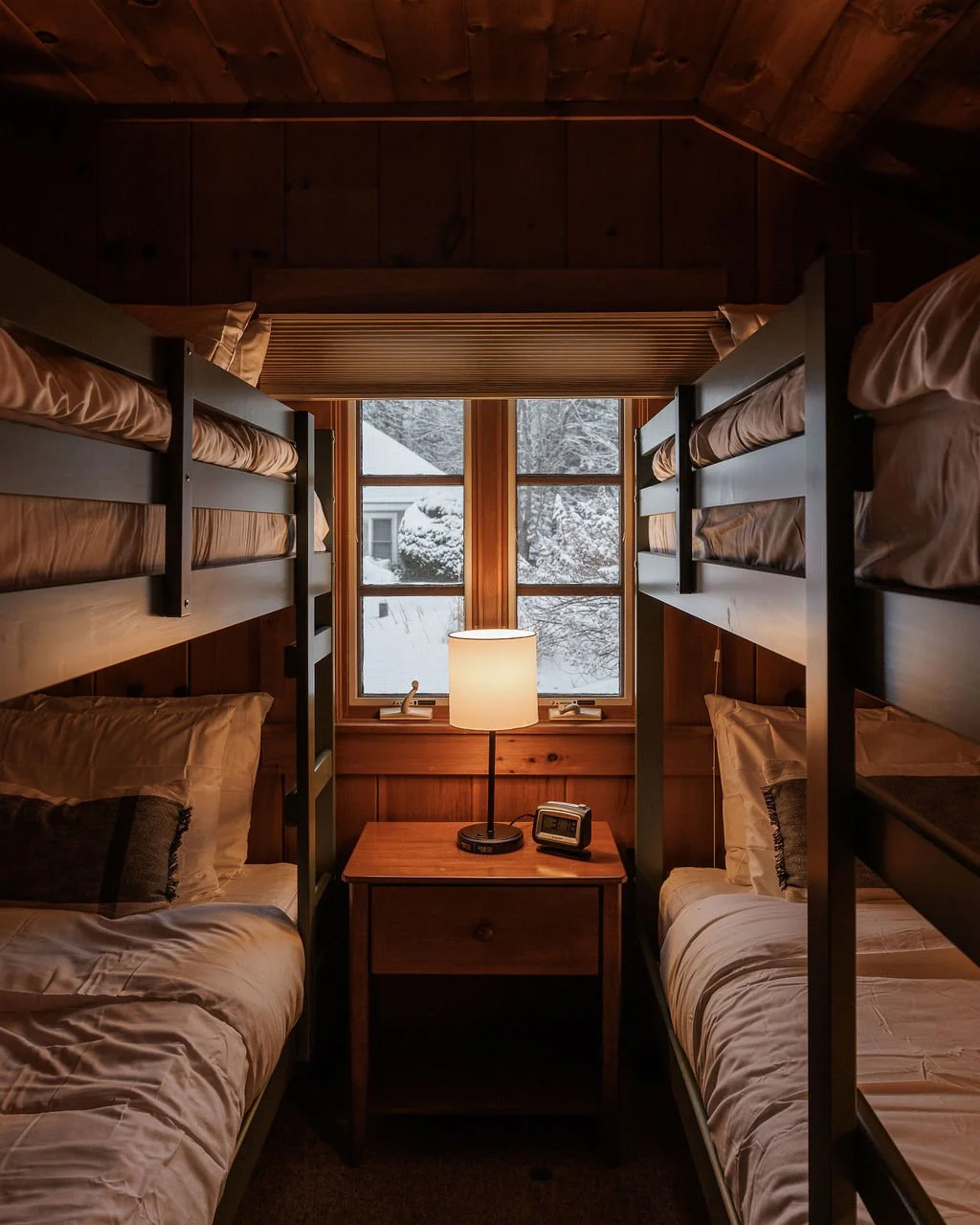 A cozy room with wooden walls and ceiling, two bunk beds with white bedding, a small wooden nightstand between them, a table lamp, and an alarm clock. A window behind the nightstand shows a snowy outdoor scene.