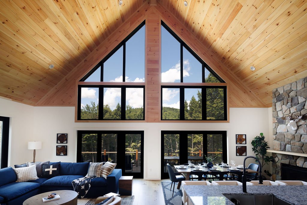 Interior of a living room with a high vaulted wooden ceiling, large windows with a view of trees and sky, a blue sectional sofa, a round coffee table, a dining table with chairs, and a stone fireplace.