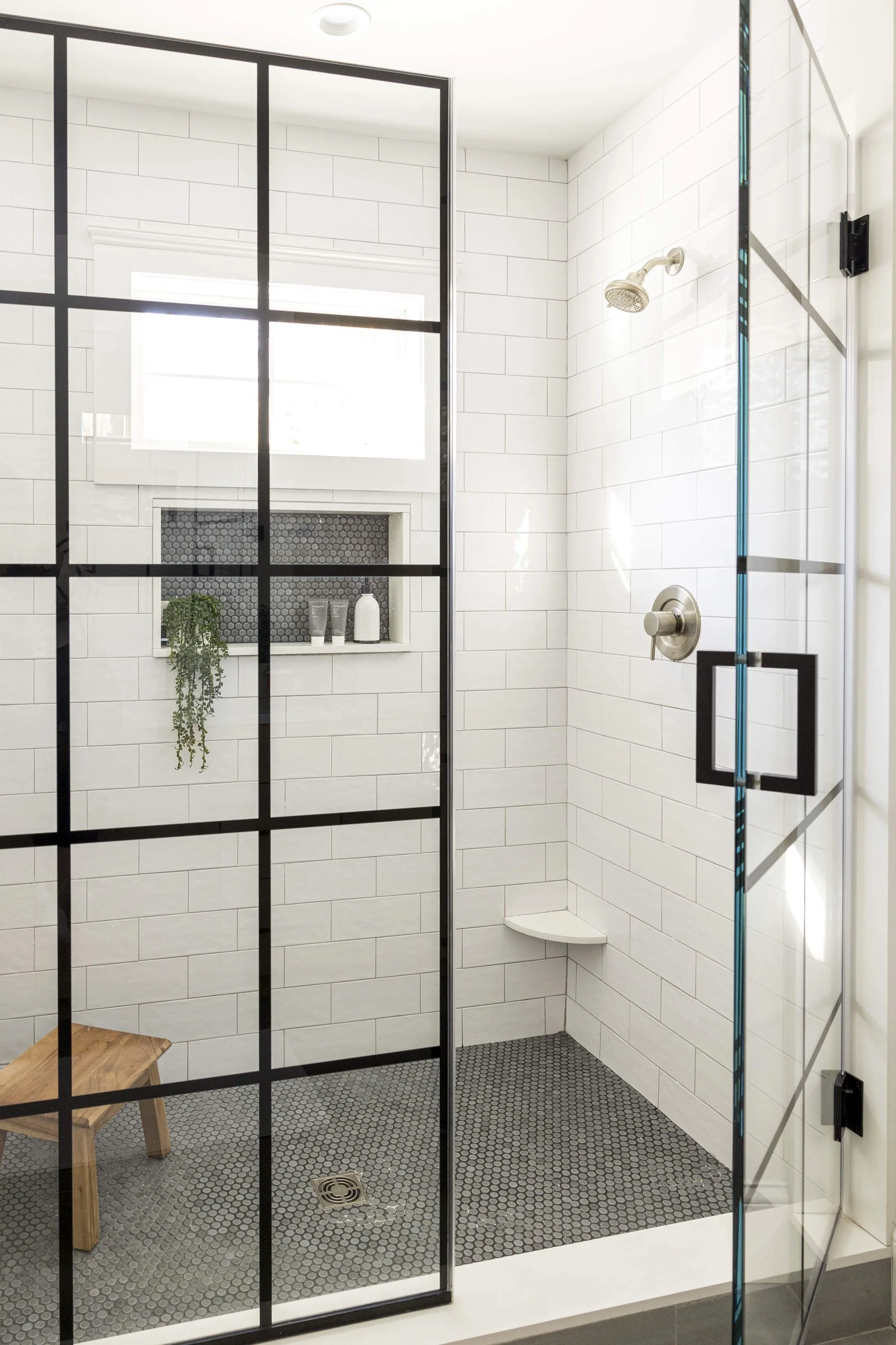 Modern walk-in shower with white subway tile walls, black metal frame glass door, small gray hexagon tile floor, a built-in shower niche with toiletries, a small white corner shower seat, a silver showerhead, a chrome shower control, a window, and a wooden bench outside the shower.