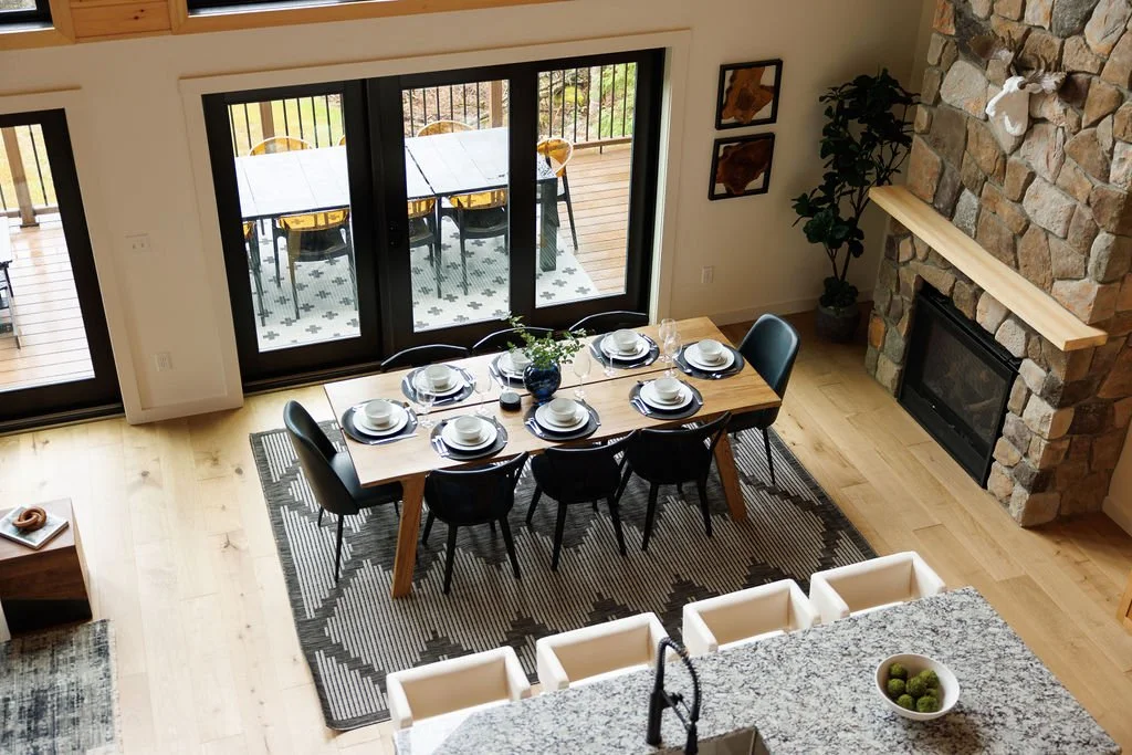 A dining area with a wooden table set for six, black chairs, and a stone fireplace. Large sliding glass doors lead to a patio with a table and chairs outside.