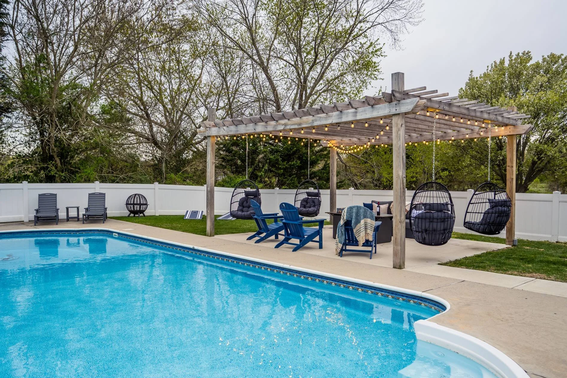 Backyard swimming pool with a white fence and a wooden pergola decorated with string lights. There are four black hanging egg chairs with cushions, two blue Adirondack chairs, and a small table with a striped towel. Two black lounge chairs are lined 