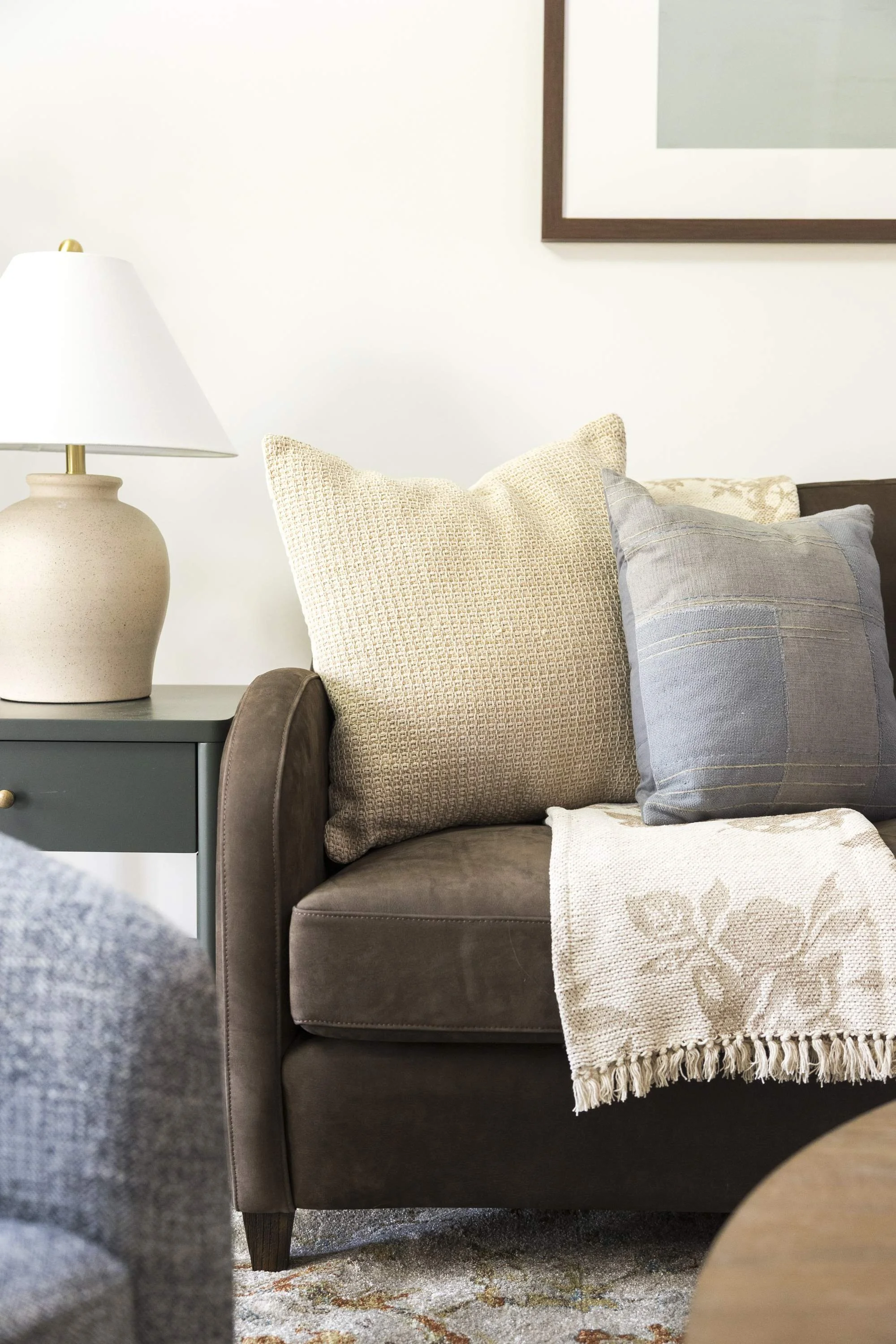 A living room corner with a brown sofa, decorative pillows, a side table with a ceramic table lamp, a framed picture on the wall, and a beige patterned throw blanket on the sofa.