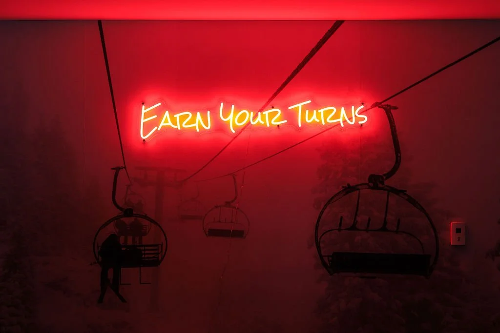 A neon sign that says 'Earn Your Turns' in red and yellow, with chairlift illustrations on a brick wall background.