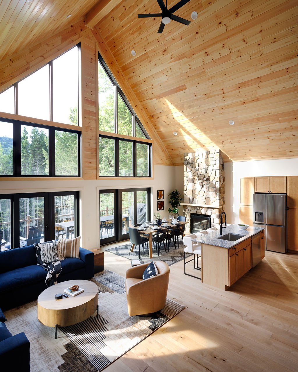 Open-concept living room and kitchen with high vaulted wooden ceiling, large windows, stone fireplace, dining area, and modern furniture.