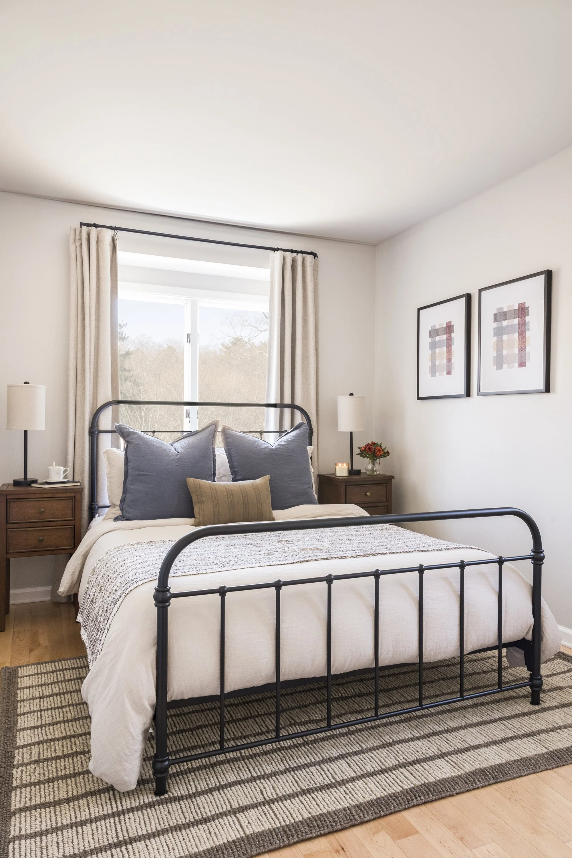 A bedroom with a black metal bed frame, beige bedding, and several pillows, including gray and beige pillows, on a striped rug. Two nightstands with lamps, a flower vase, and a candle are on each side of the bed. A window with beige curtains is behin