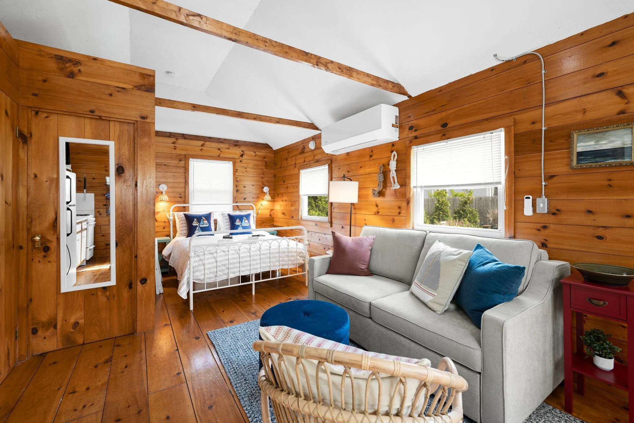 A cozy bedroom with wooden walls and ceiling beams, featuring a white metal bed with nautical-themed pillows, a white sofa with colorful pillows, and a small wicker chair, illuminated by natural light through multiple windows.