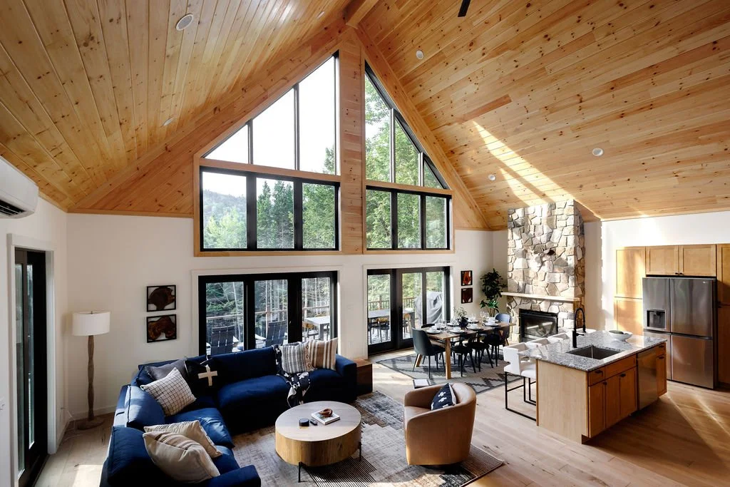 Living room with high wooden vaulted ceiling, large windows, stone fireplace, dining table, kitchen island, and modern furniture.