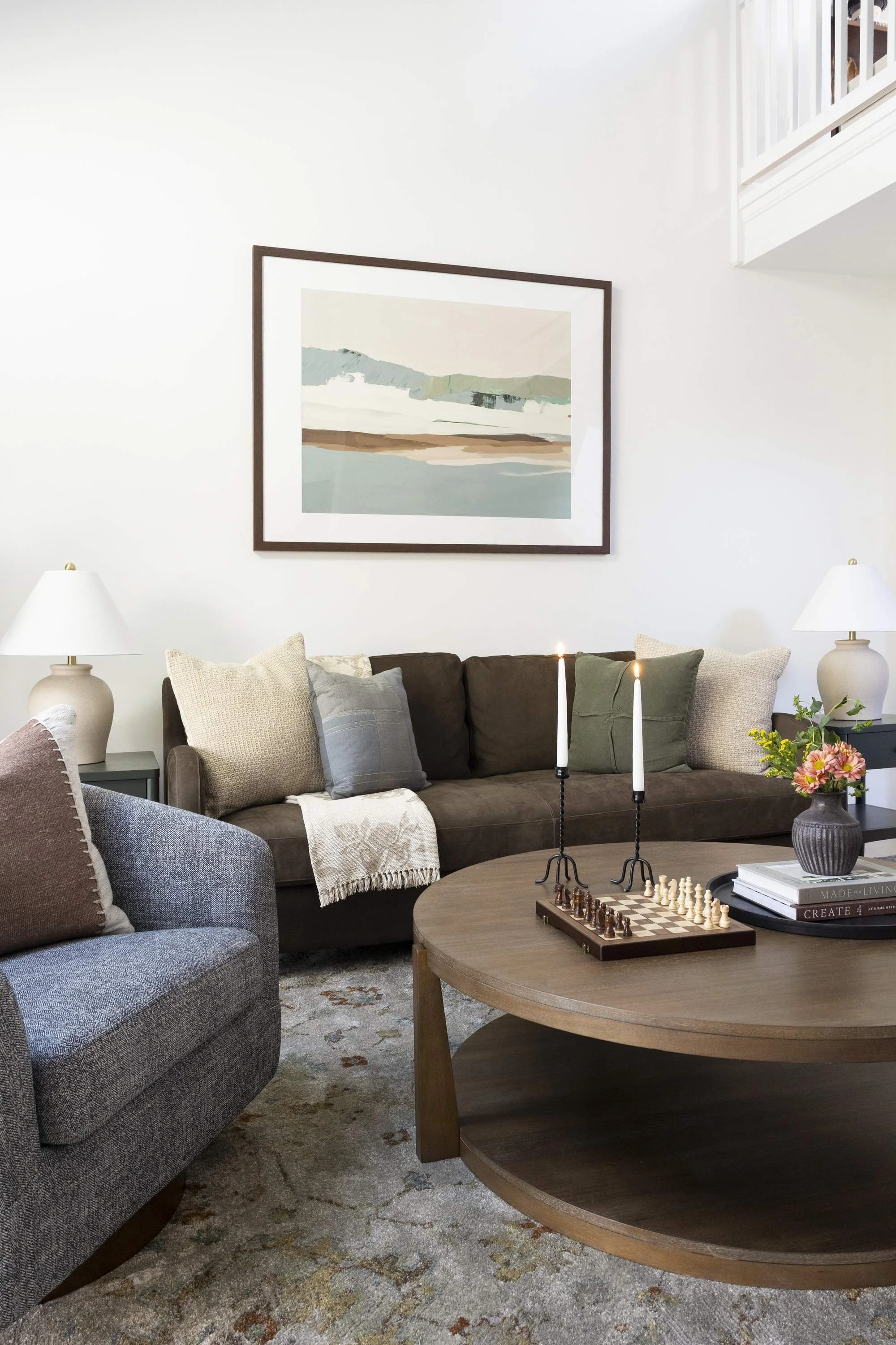 A living room with a dark brown sofa decorated with beige, gray, and green pillows. Behind the sofa is a white wall with a large framed abstract landscape painting. On the right, a side table with a beige lamp with a white lampshade. In the foregroun