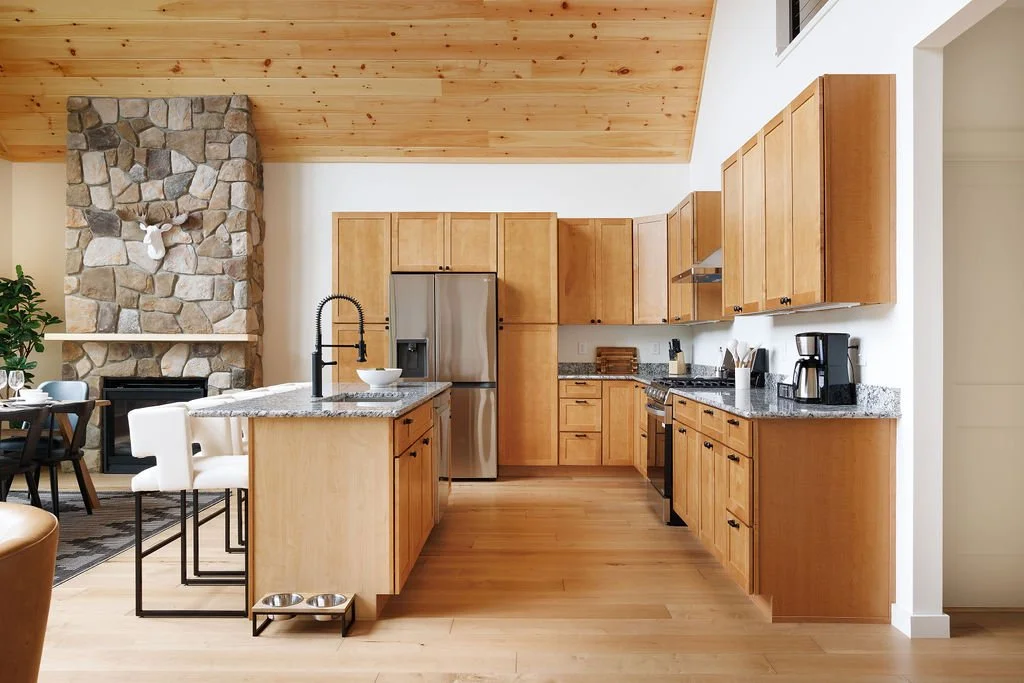 Kitchen with wooden cabinets, granite countertops, stainless steel refrigerator, black oven, coffee maker, and a stone fireplace with a mounted deer head, in a modern open-concept home.