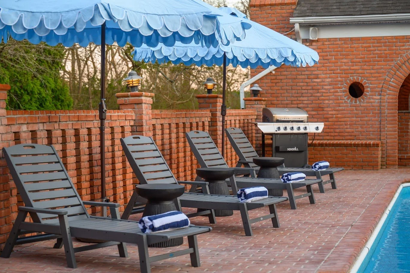 Row of four gray lounge chairs with rolled towels on the footrests, arranged on a brick poolside patio, under a blue patio umbrella, with a brick wall and gas grill in the background.