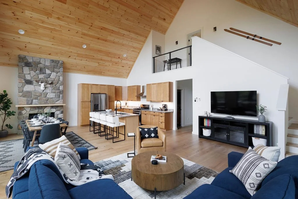 Open-concept living room and kitchen area in a modern home with a vaulted wooden ceiling, stone fireplace, blue sofa, dining table, kitchen island, and TV on a black stand.