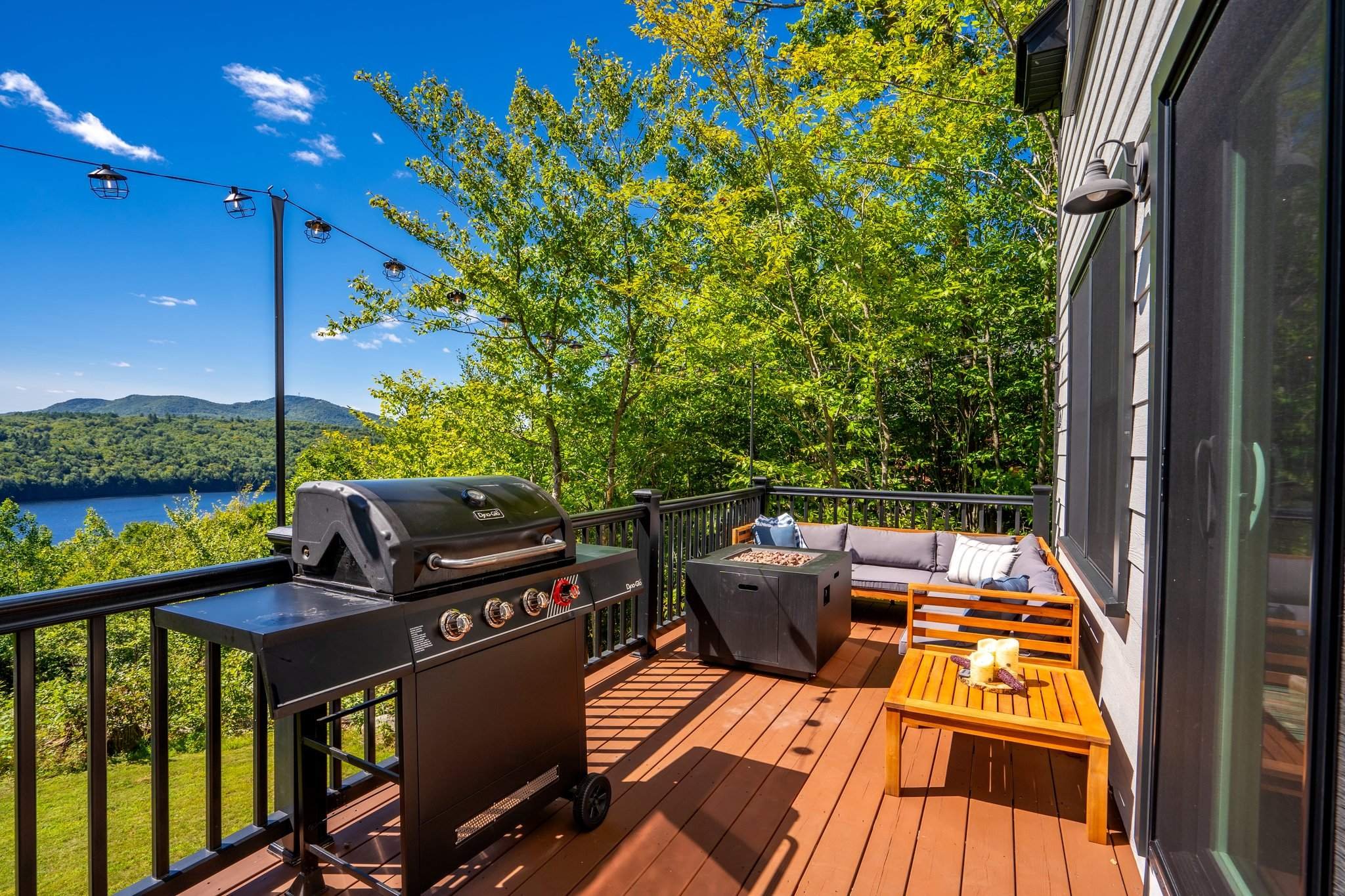 A balcony overlooking a scenic view of a lake and green mountains. The balcony has a gas grill, a cushioned outdoor sofa, a wooden bench, candles, and string lights hanging above.