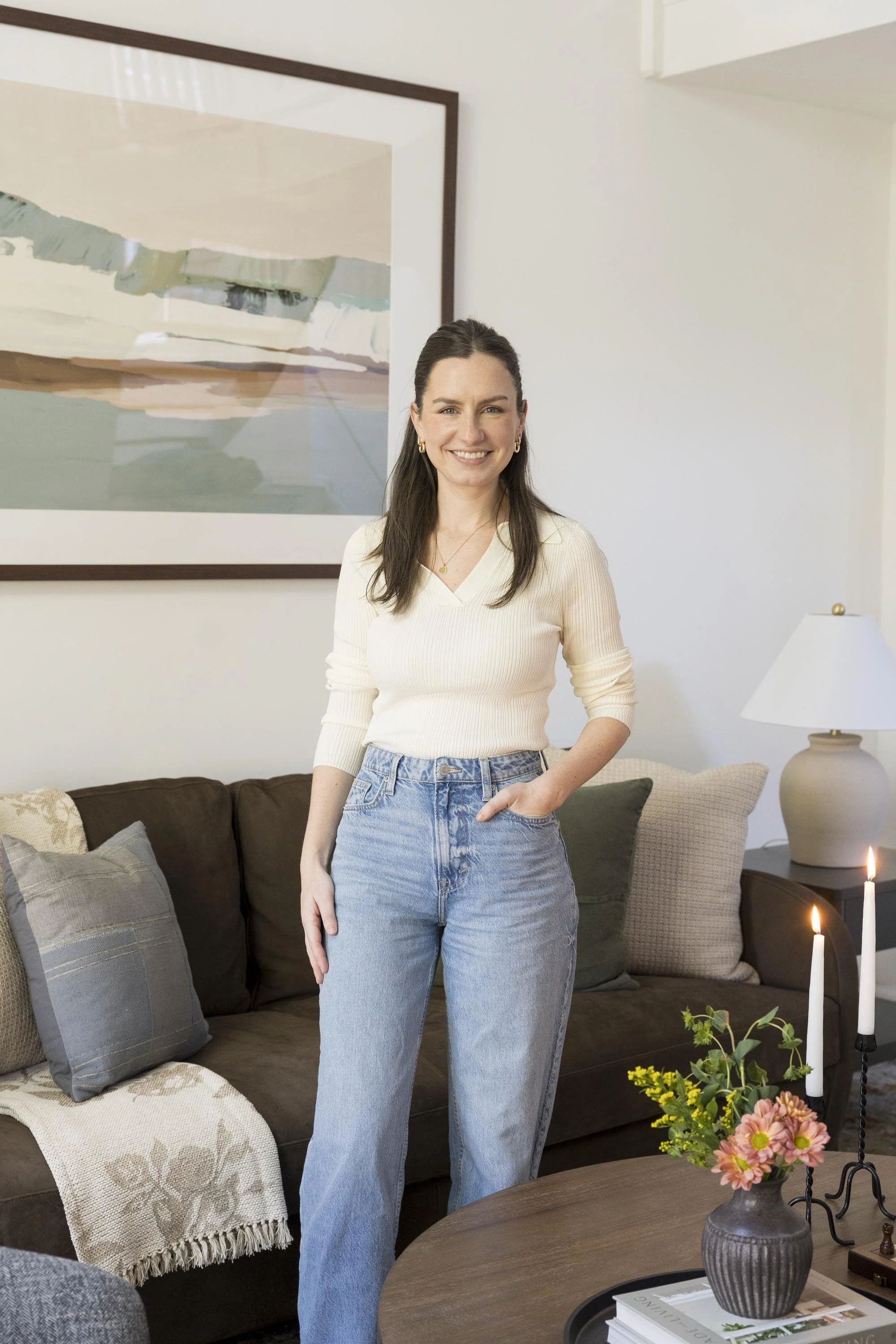 A woman with long dark hair wearing a cream-colored ribbed sweater and high-waisted jeans stands in a living room, smiling. Behind her, there's a brown couch with various pillows, a large abstract painting on the wall, and a side table with a white l