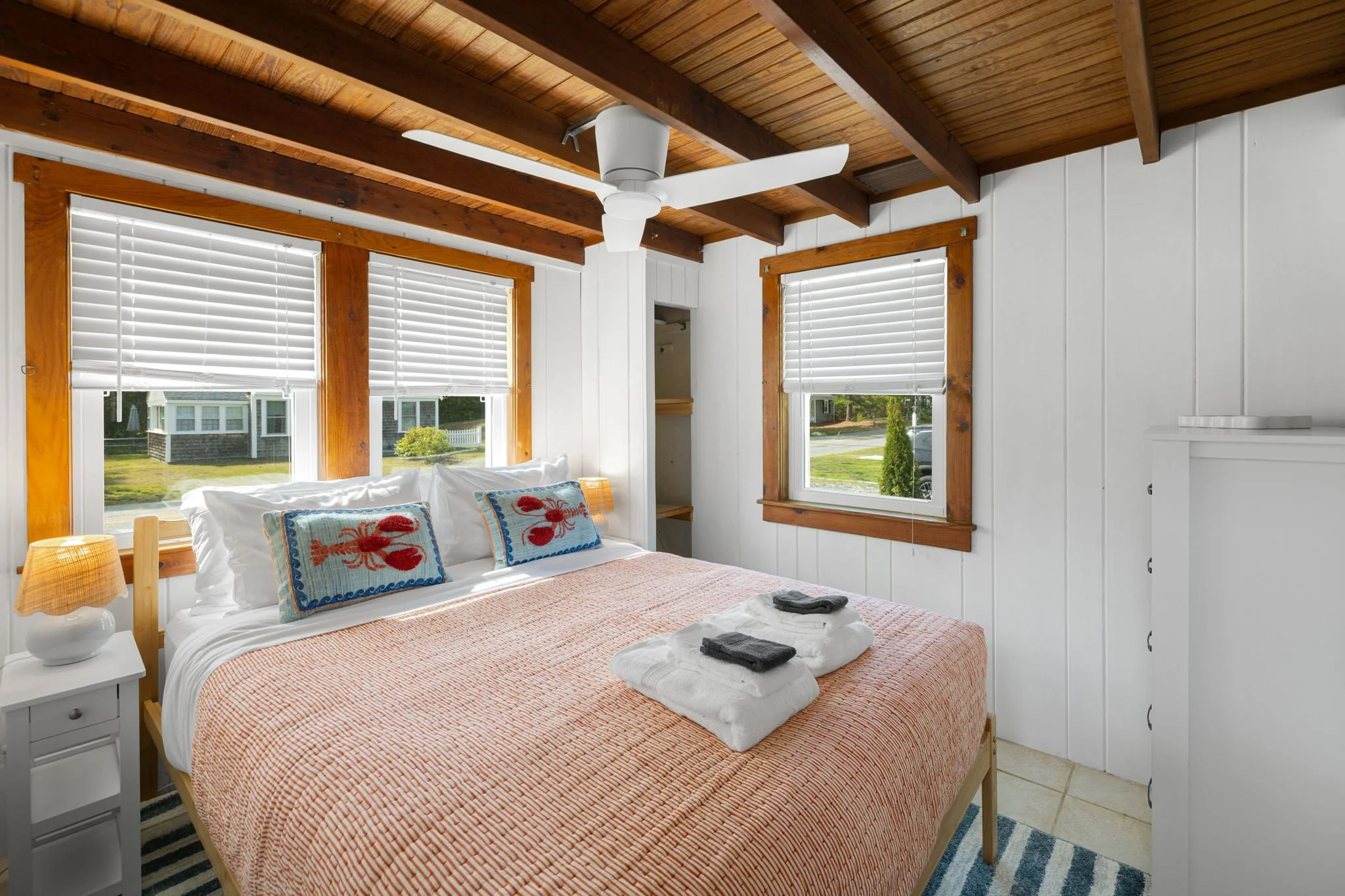 Cozy bedroom with white walls, two windows with blinds, a bed with an orange and white striped bedspread, lobster decorative pillows, and folded towels on the bed. Wooden ceiling beams and ceiling fan. Nightstand with lamp on the left.