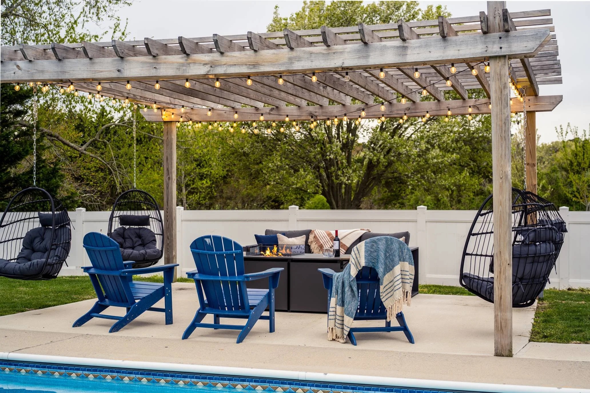 A backyard patio with a wooden pergola decorated with string lights. Blue and black lounge chairs, a small fire pit table with a fire burning, and outdoor cushions are arranged around the table. There are trees and a white fence in the background.