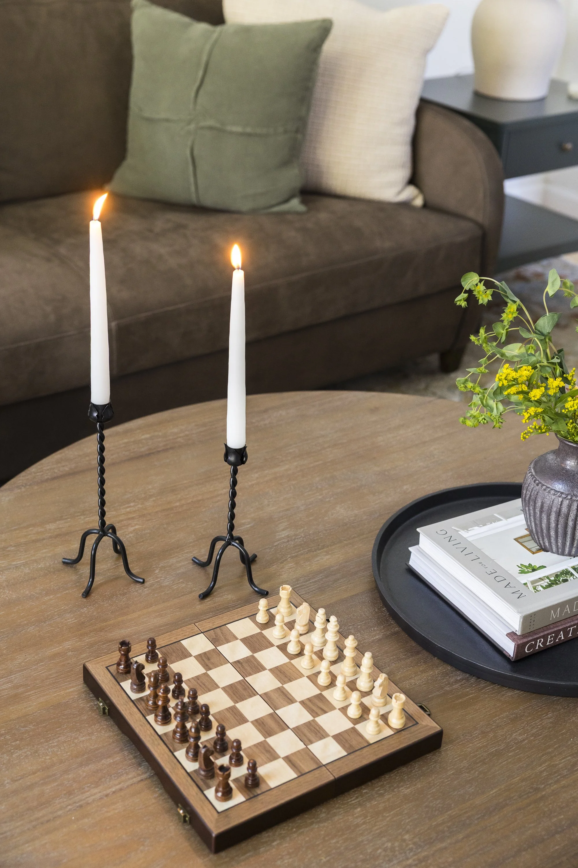 A wooden chessboard with chess pieces set up for a game is on a wooden table. Two black lit candles in twisted black candle holders are nearby. On the right, there is a stack of books on a black tray, with a gray vase holding green leafy plant on top