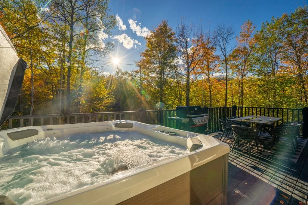 Hot tub on an outdoor deck with a table and chairs, a barbecue grill, surrounded by trees with autumn foliage under a clear blue sky.