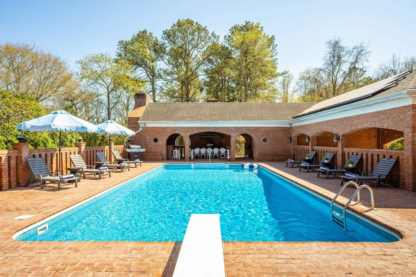  residential backyard swimming pool with lounge chairs, umbrellas, and a brick house with a covered patio, surrounded by trees.