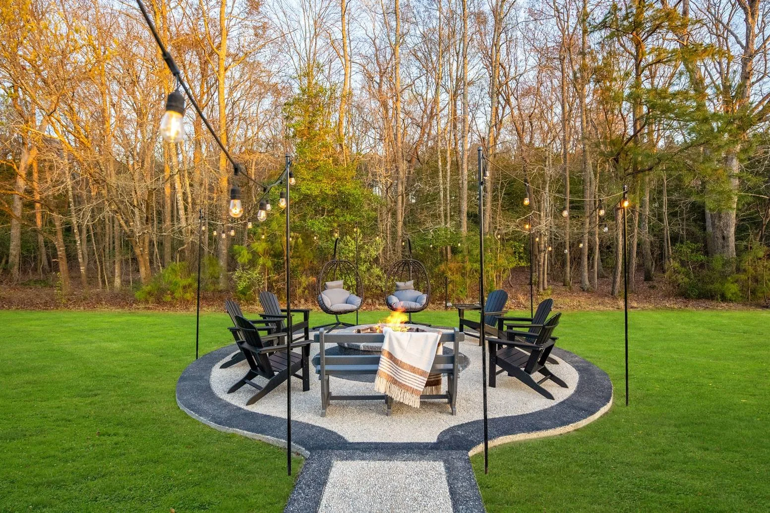 Outdoor wedding or event setup with chairs arranged around a fire pit, hanging string lights, and hanging chairs, on a landscaped lawn with a wooded background at sunset.