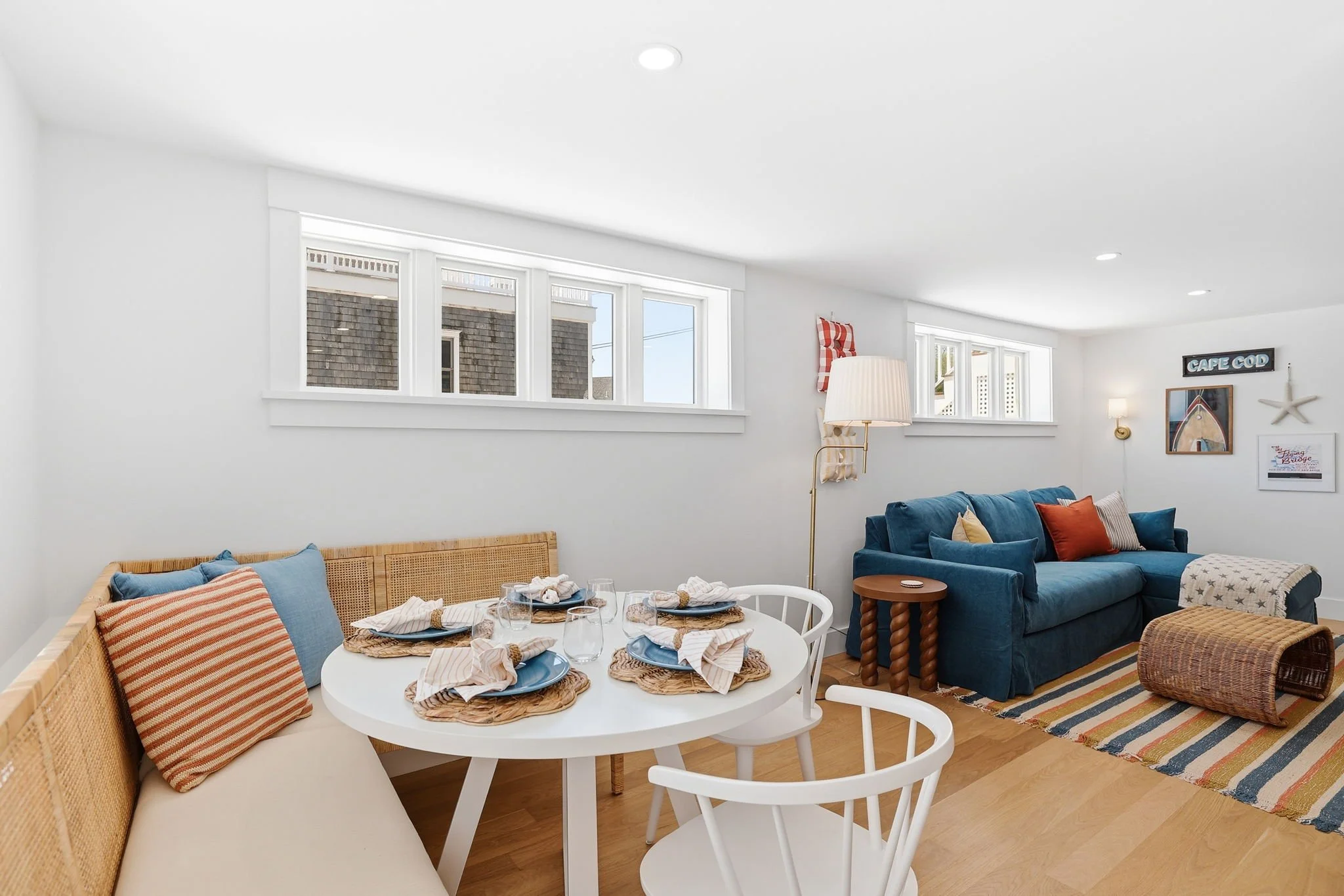 Cozy living room with white walls and ceiling, windows near the ceiling, a blue sofa with orange and beige pillows, a wooden side table, a striped rug, a dining table with chairs set for a meal, and wall decorations including a sign reading "CAPE COD