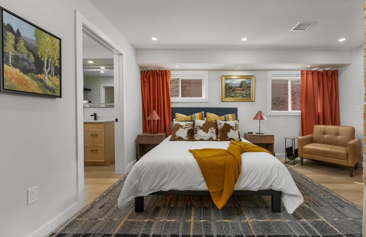 A bedroom with white walls, hardwood flooring, a bed with white bedding and multiple patterned pillows, orange curtains, a tan armchair, two nightstands with pink lamps, and framed landscape paintings on the wall.