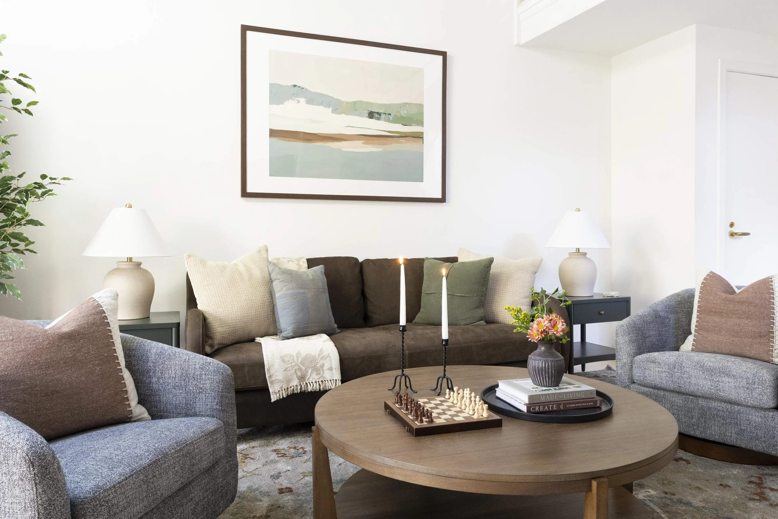 A cozy living room with a brown sofa, gray and beige armchairs, and a round wooden coffee table with a chessboard and books. There are two white table lamps, a vase of flowers, and a large abstract landscape painting on the white wall.