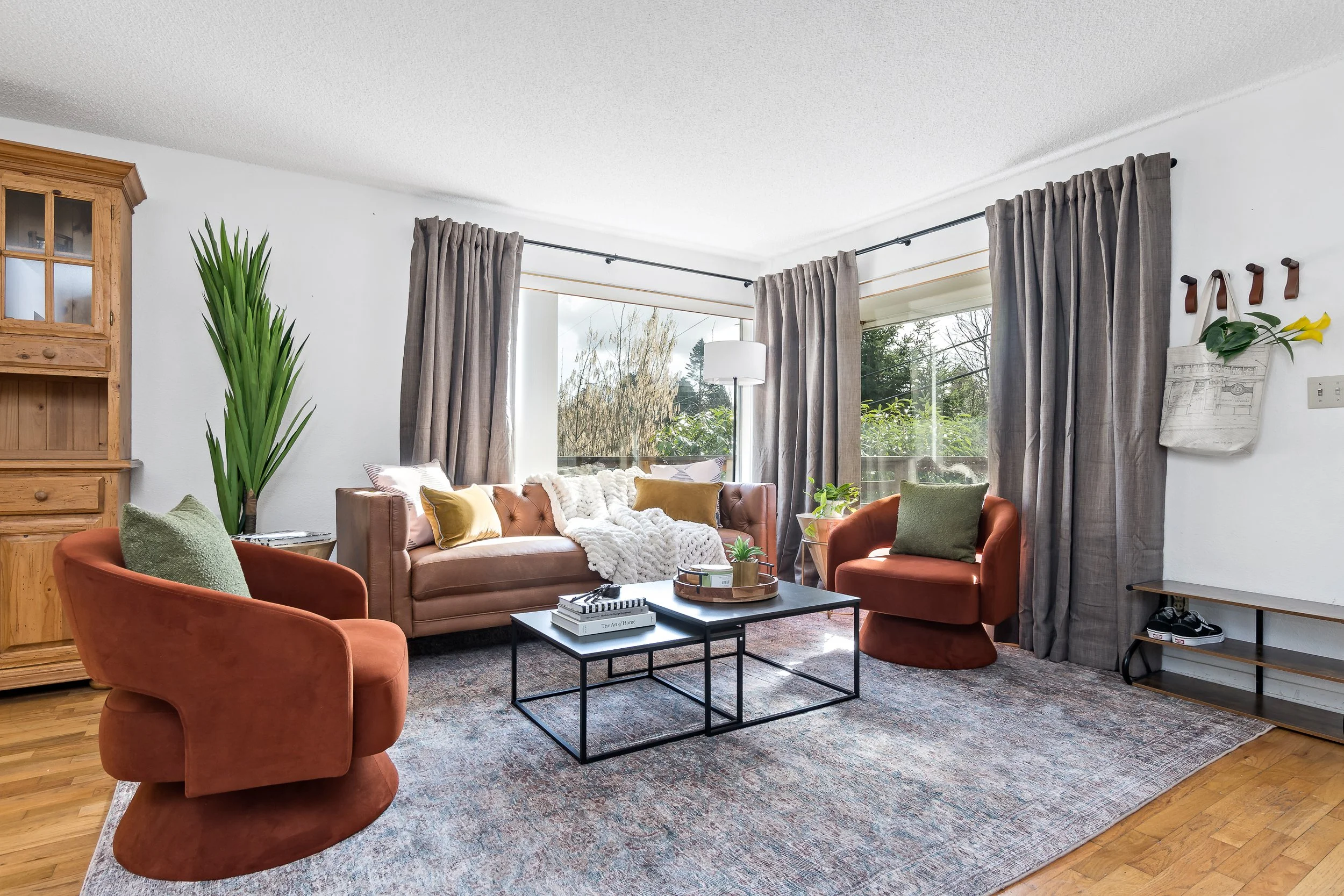 Living room with a pink sofa, orange chairs, a black coffee table, and large windows with beige curtains, decorated with a wall-mounted bag and flower hanging on the wall.