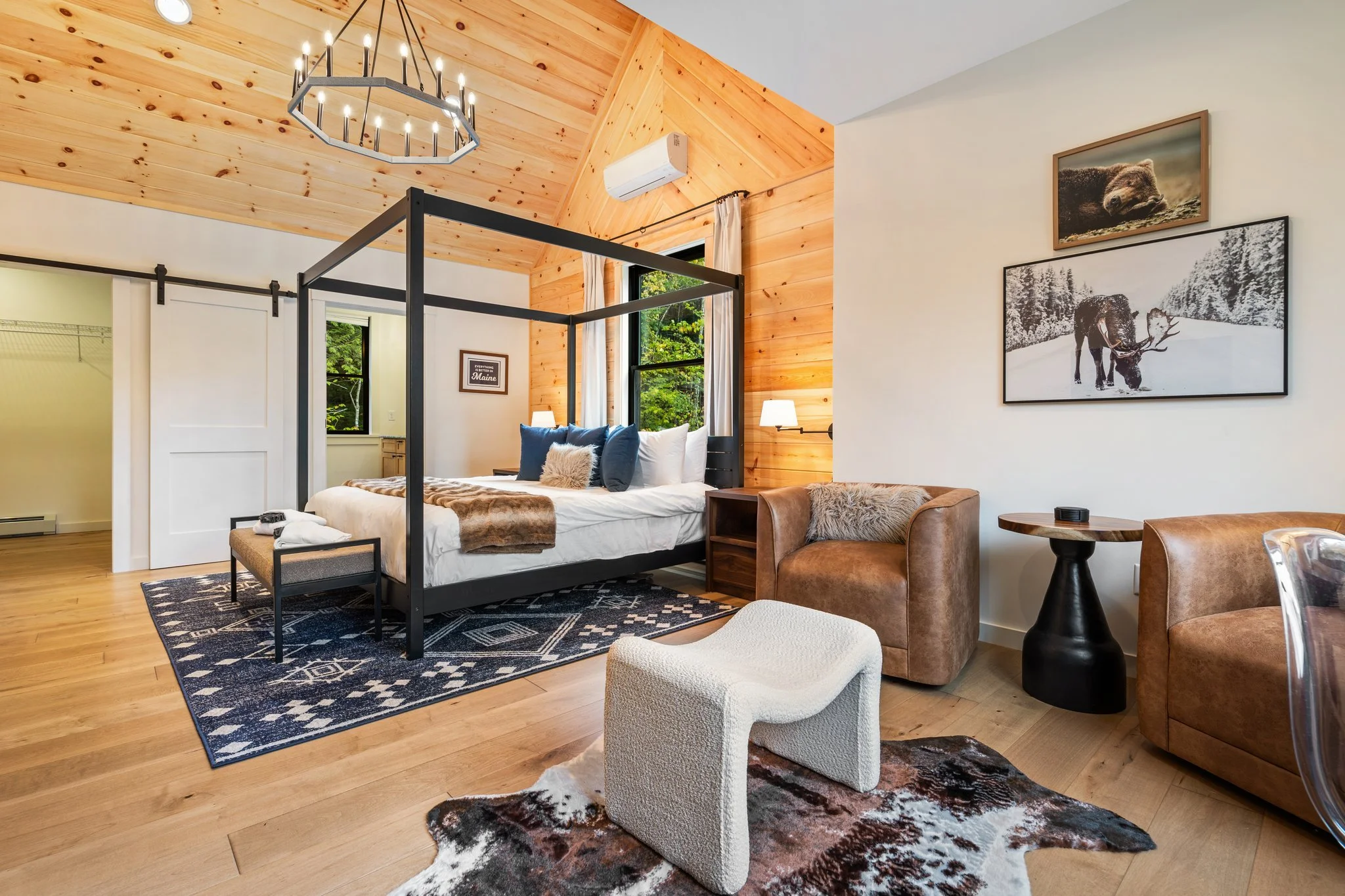 A bedroom with a vaulted wood ceiling, a blackout canopy bed with navy and white pillows, a cozy throw, and a rug with a geometric pattern. There are framed pictures of animals on the wall, a wooden side table, brown leather armchair, an ottoman, a c