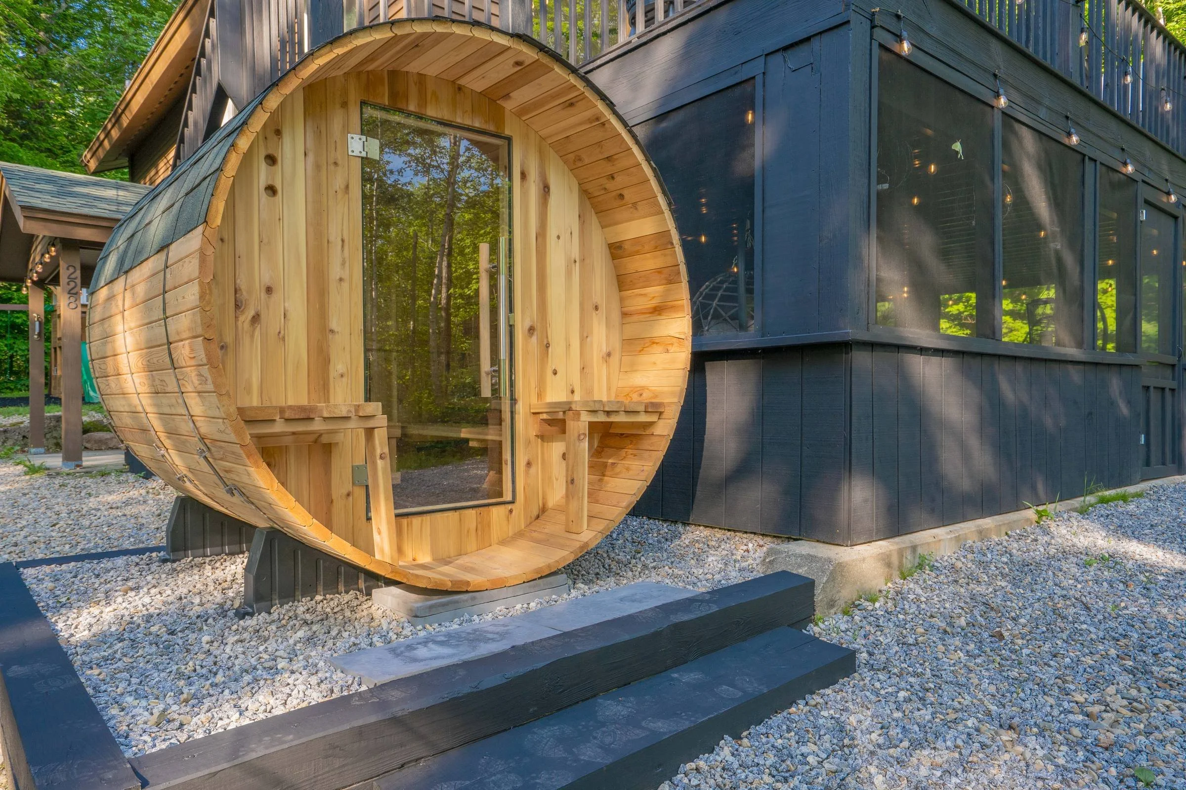 A modern tiny house with a curved wooden front and large glass door, nestled among green trees, with string lights hanging from the upper deck.