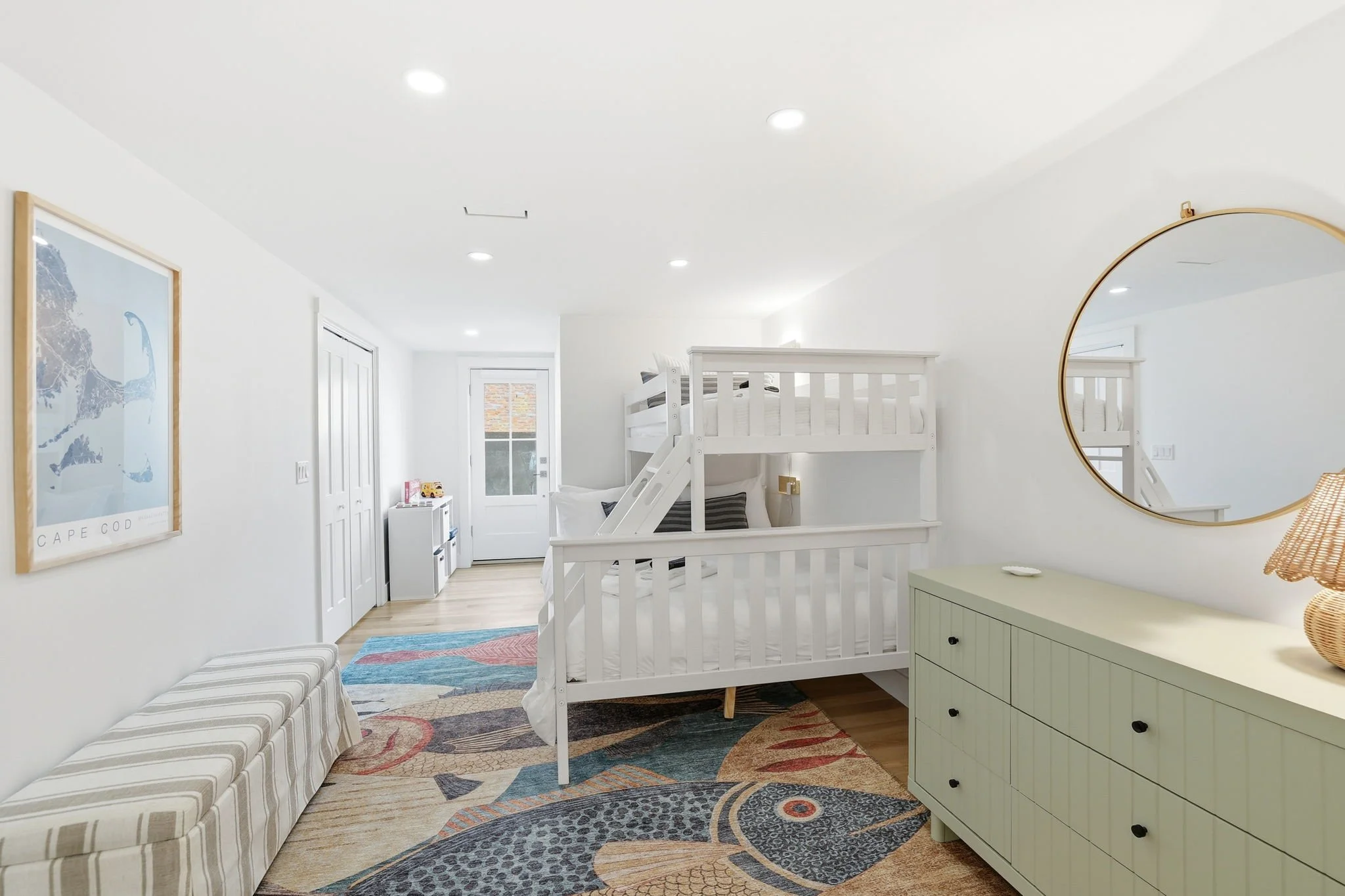 A bedroom with white walls and ceiling, including a loft bed with black and white bedding, a striped bench, a green dresser, a round mirror, and framed artwork on the wall. There are recessed lights in the ceiling and a colorful area rug on the woode