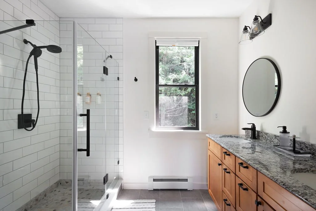 Modern bathroom with a glass-enclosed shower, white subway tiles, a granite countertop vanity, a round mirror, and a window showing greenery outside.