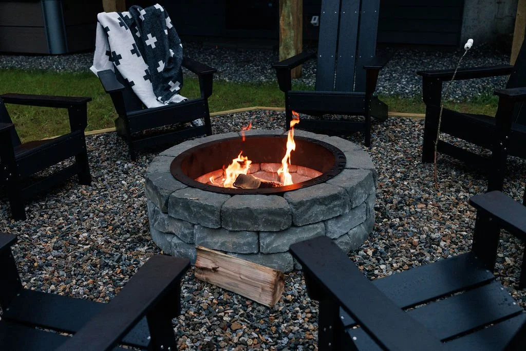 A circular firepit made of gray stone bricks surrounded by six black chairs on a gravel surface outdoors with a wooden house and grass in the background.