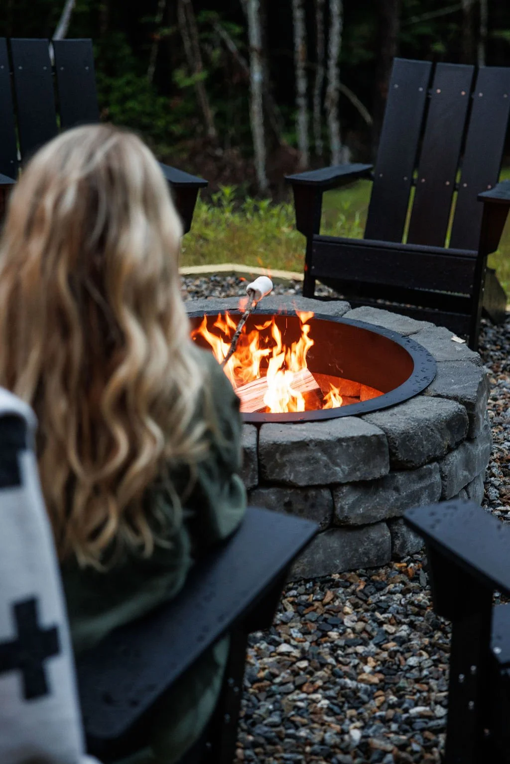 A person with long, wavy blonde hair roasting marshmallow over a campfire fire pit surrounded by black wooden Adirondack chairs in a wooded setting.