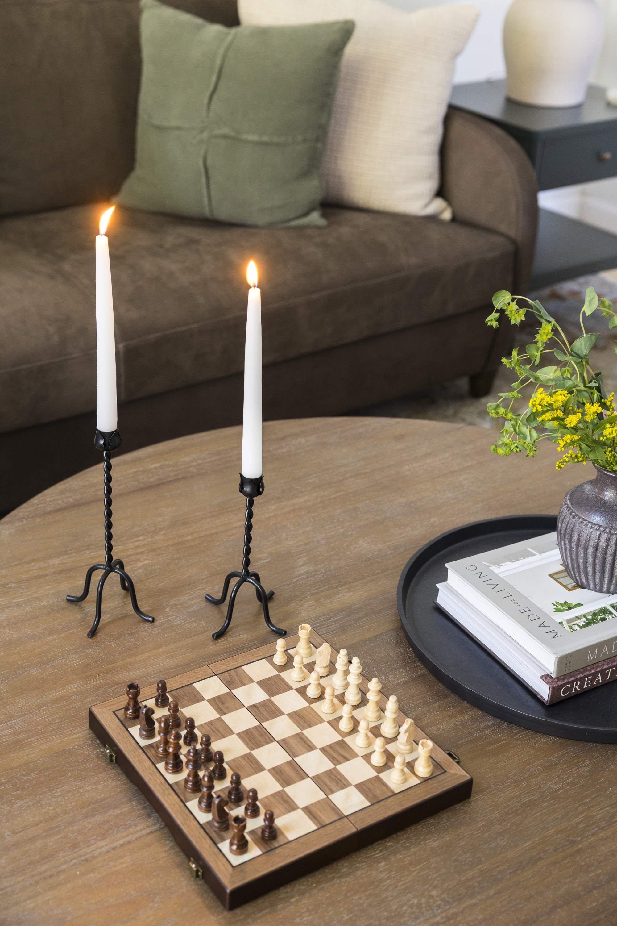 A chessboard and pieces on a wooden table, with two white candles in black candle holders, a vase with flowers, books, and a sofa with cushions in the background.